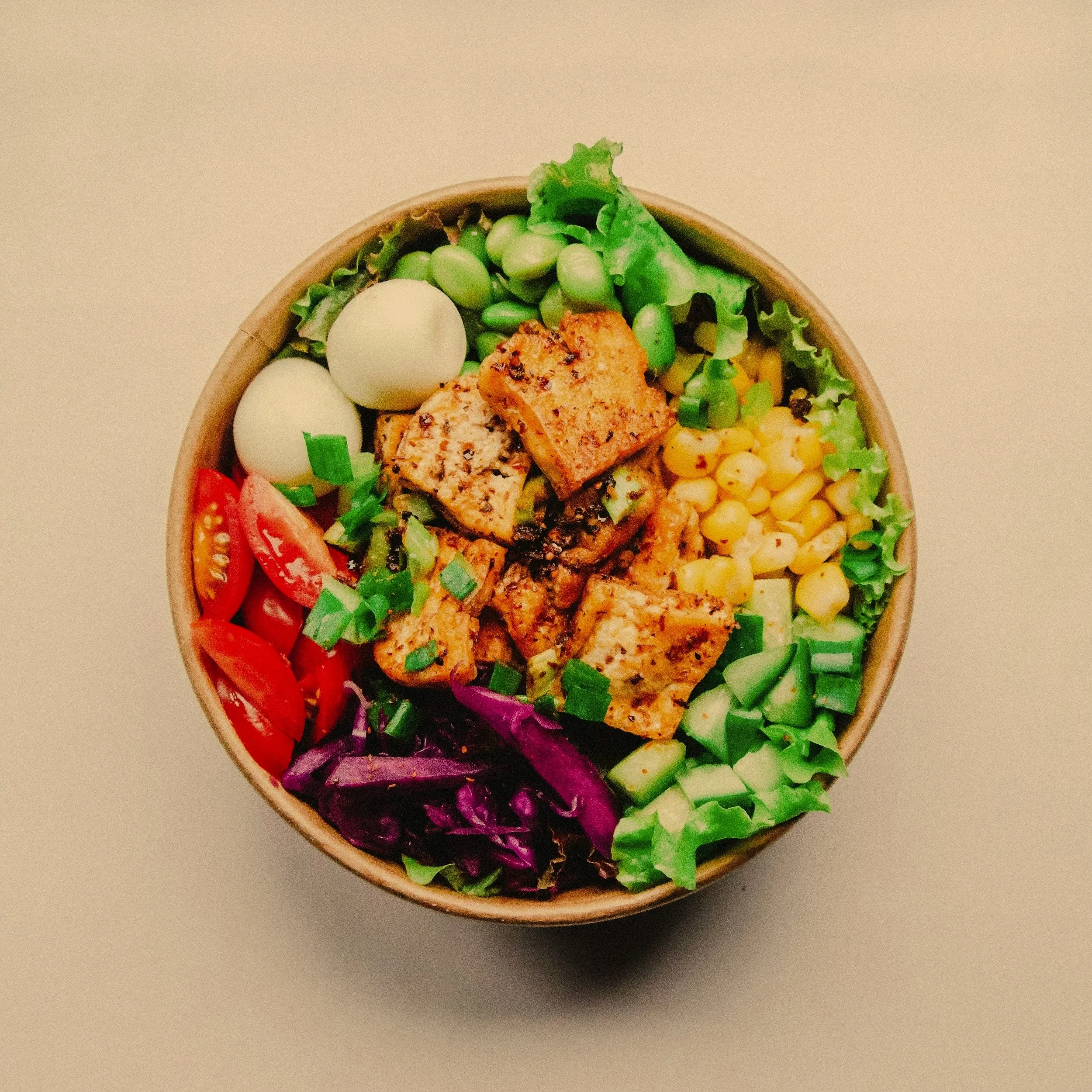 Close-up of a bowl filled with various nutritious fresh vegetables including cherry tomatoes, corn, greens, cucumbers, and slices of seasoned grilled chicken, on a plain background.