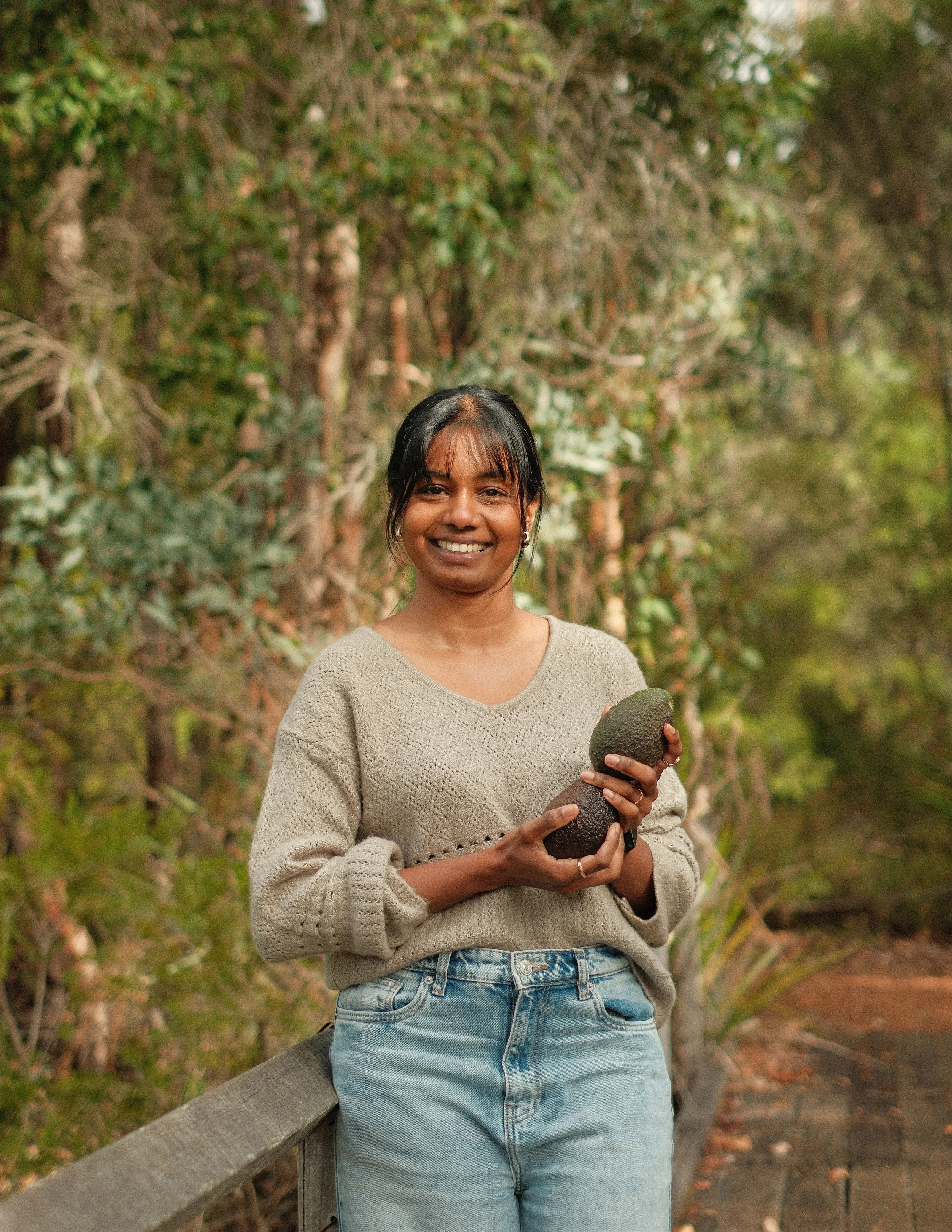 A smiling woman holds an avocado while standing outdoors in a nature setting.
