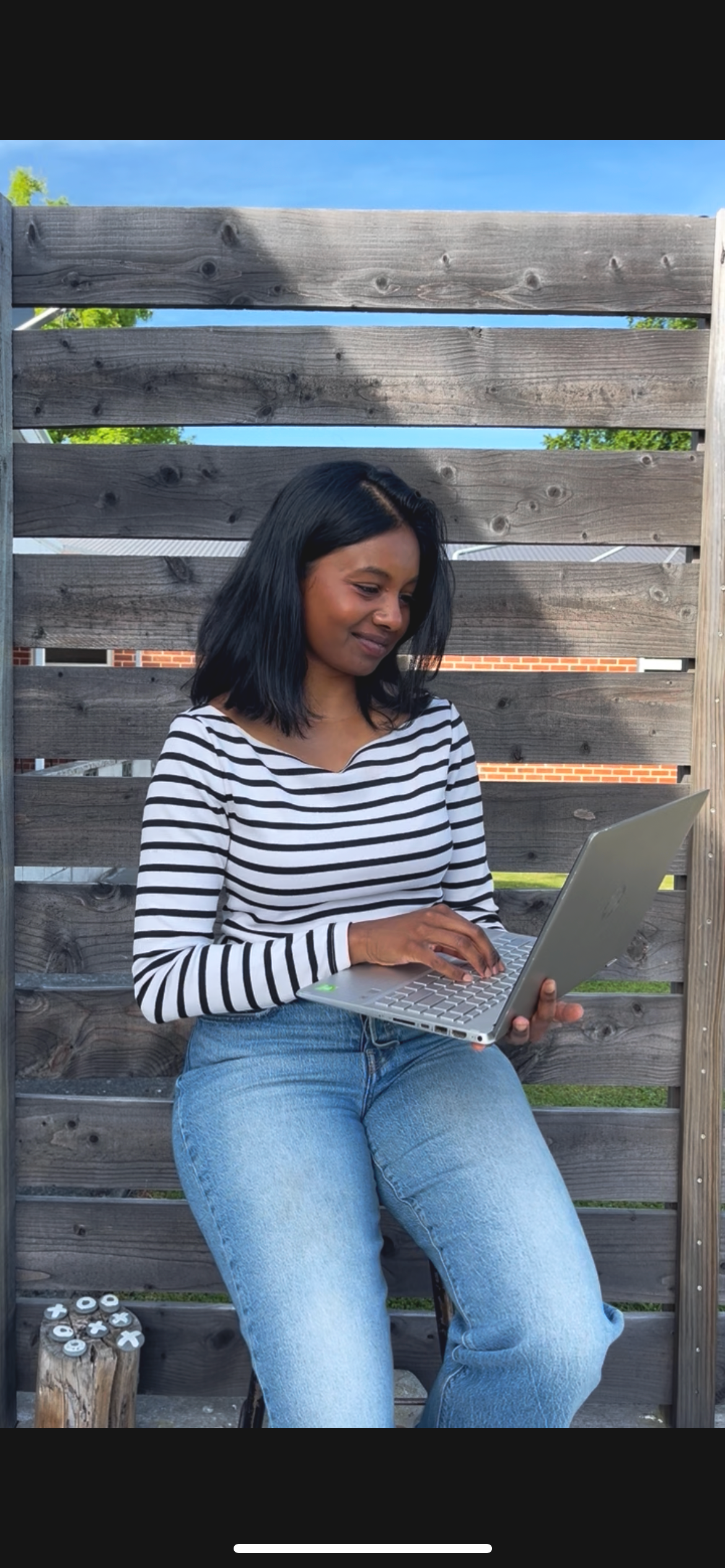 A woman with shoulder-length black hair, wearing a black and white striped long-sleeve shirt and blue jeans, sitting outside and using a silver laptop. She is sitting against a wooden privacy fence with a blue sky and a brick building in the background.