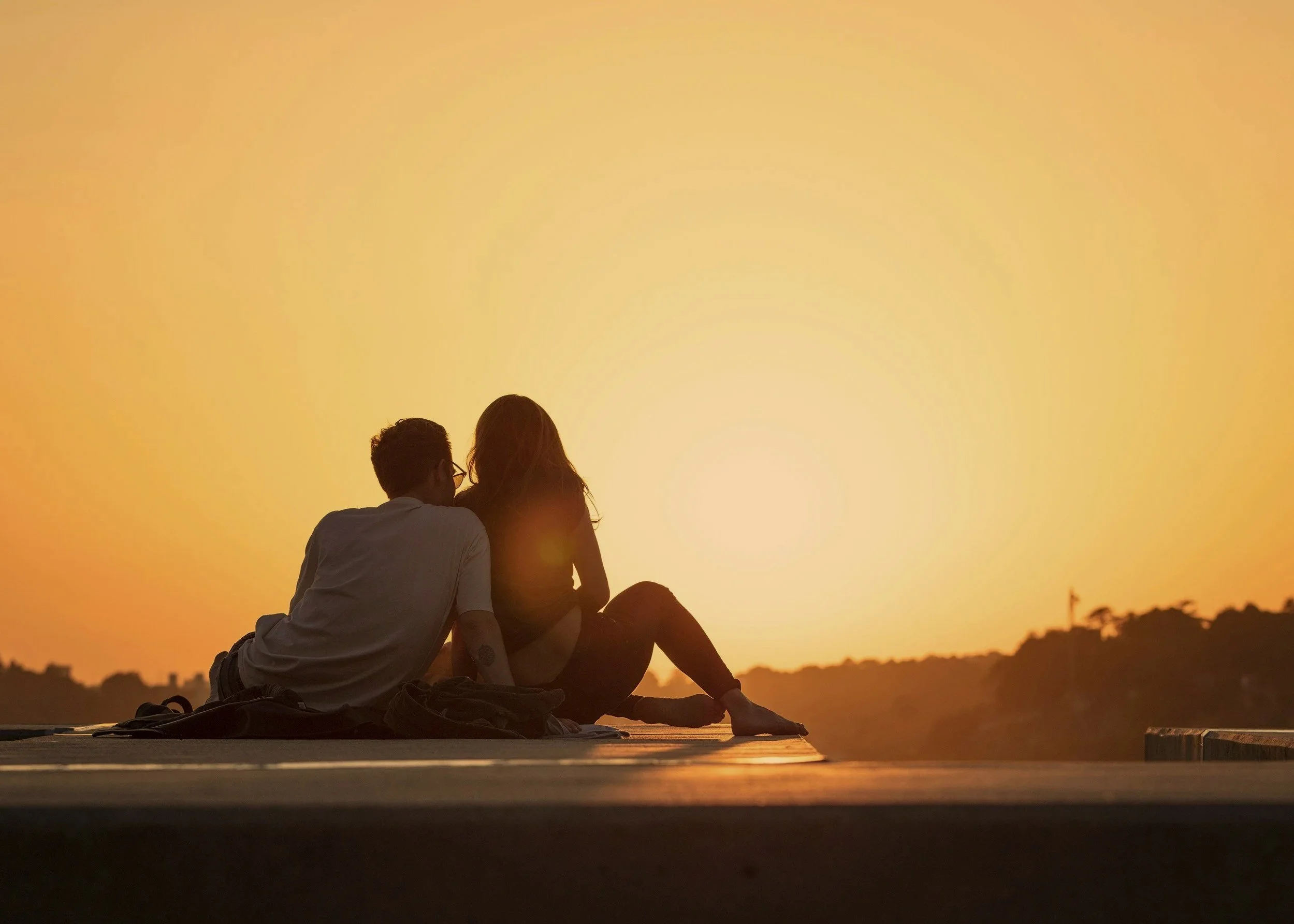 Two people sitting close together by a body of water during sunset, with a warm orange sky in the background.