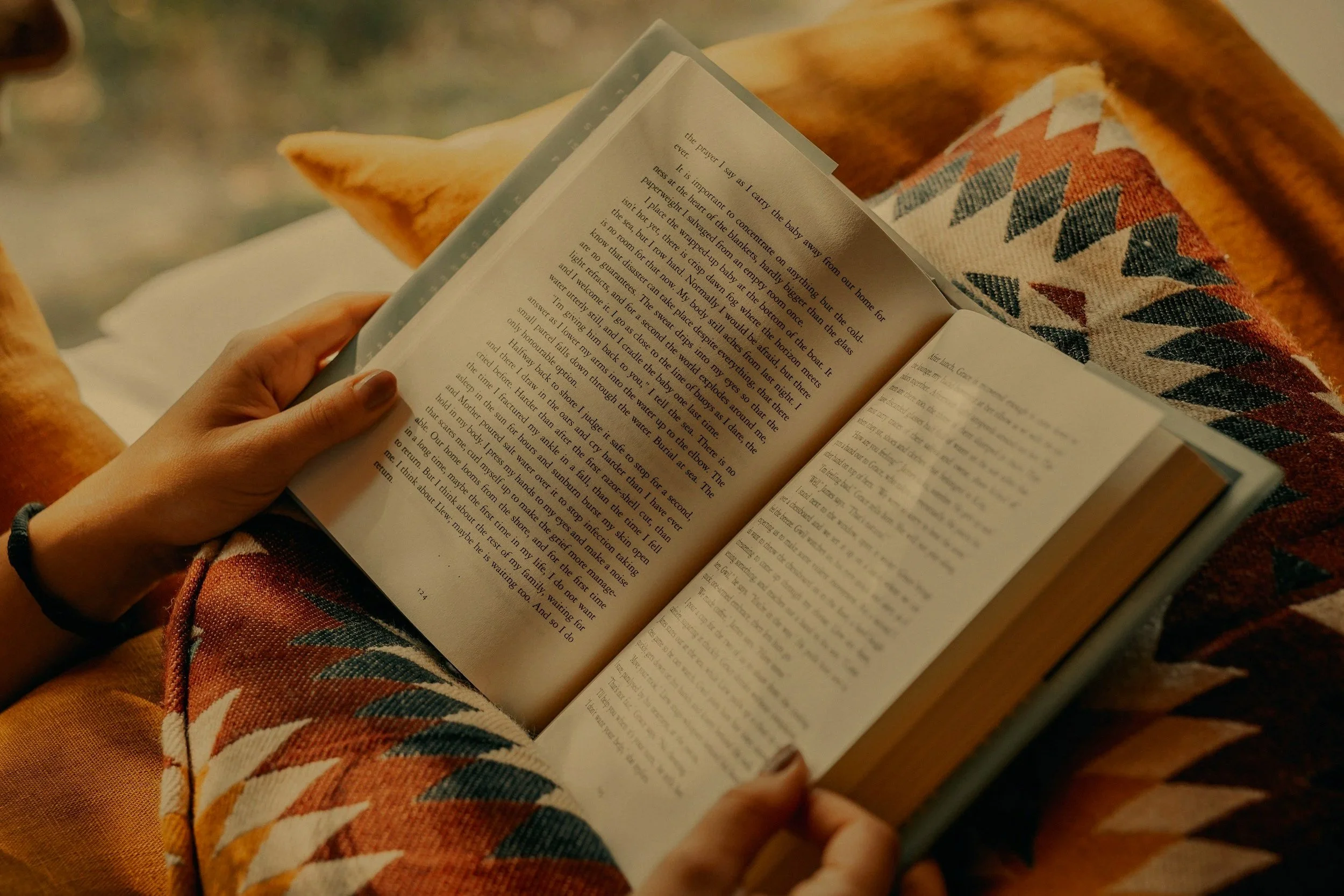 A person wearing a patterned long-sleeve shirt, sitting on a piece of furniture, is reading a book. The focus is on their hands and the open book, with a blanket or cushion with a geometric design partially visible.
