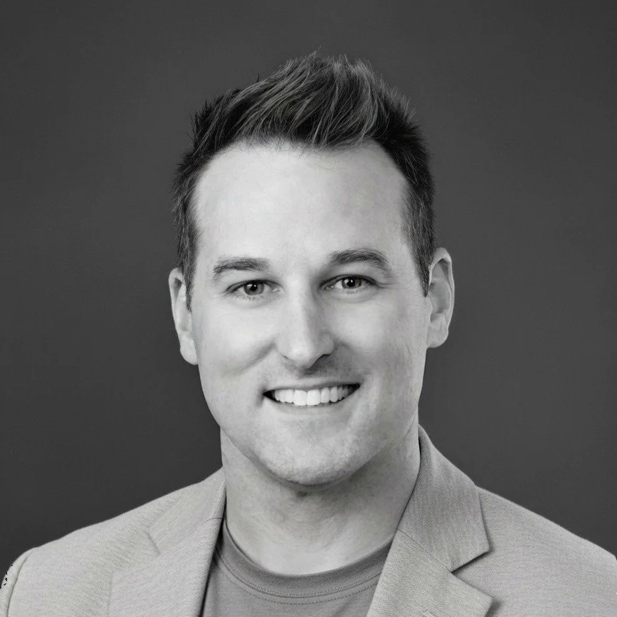 Black and white portrait of a smiling man with short, styled hair, wearing a blazer over a shirt, against a plain background.