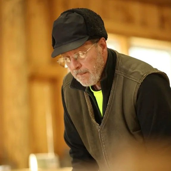 A man with glasses, a beard, and gray hair wearing a black cap, green vest, and black shirt, looking down indoors with wooden walls in the background.