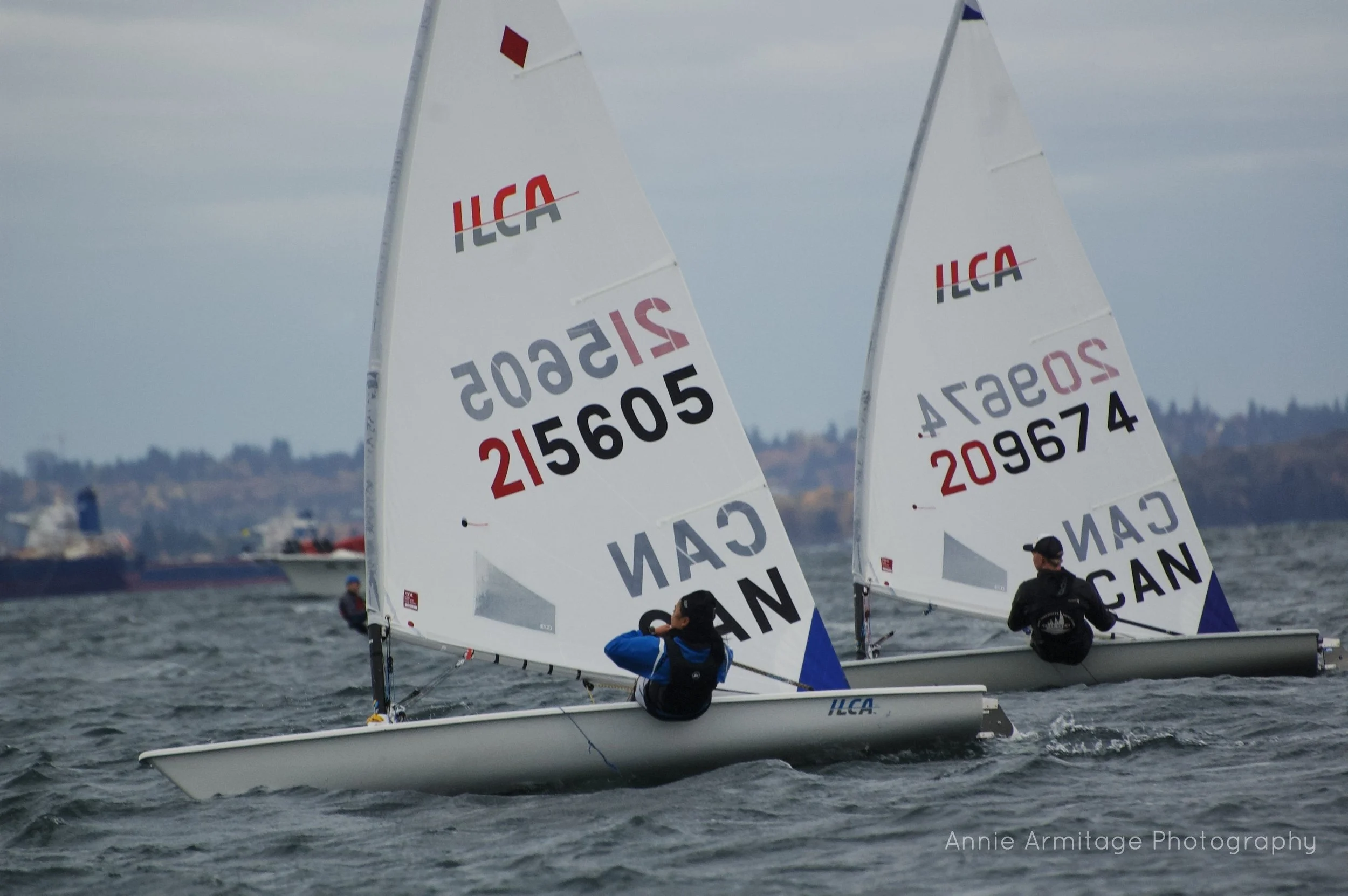 Two sailors racing in small sailboats on a body of water with a city skyline in the distance. The boats are marked with ILCA and have sail numbers. The sailors are dressed in gear suitable for sailing.