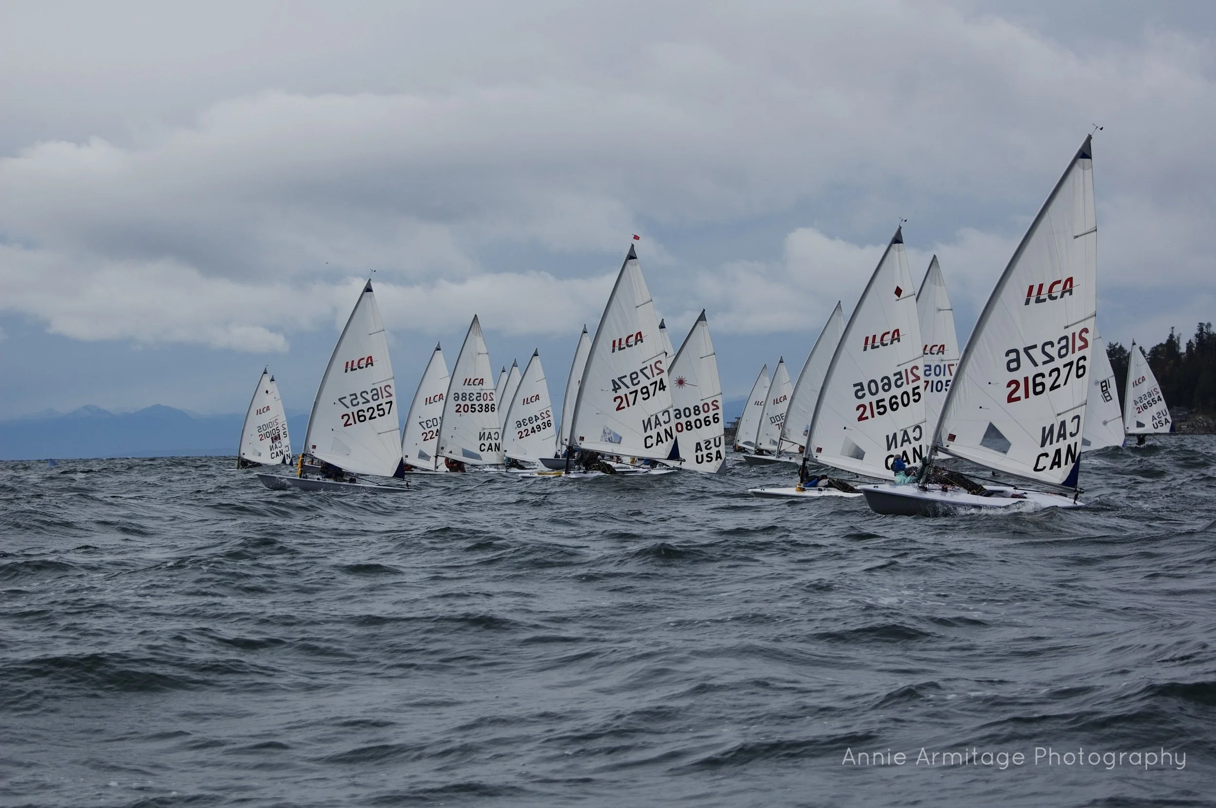 Multiple sailboats racing in choppy waters under cloudy sky, with mountains visible in the background.