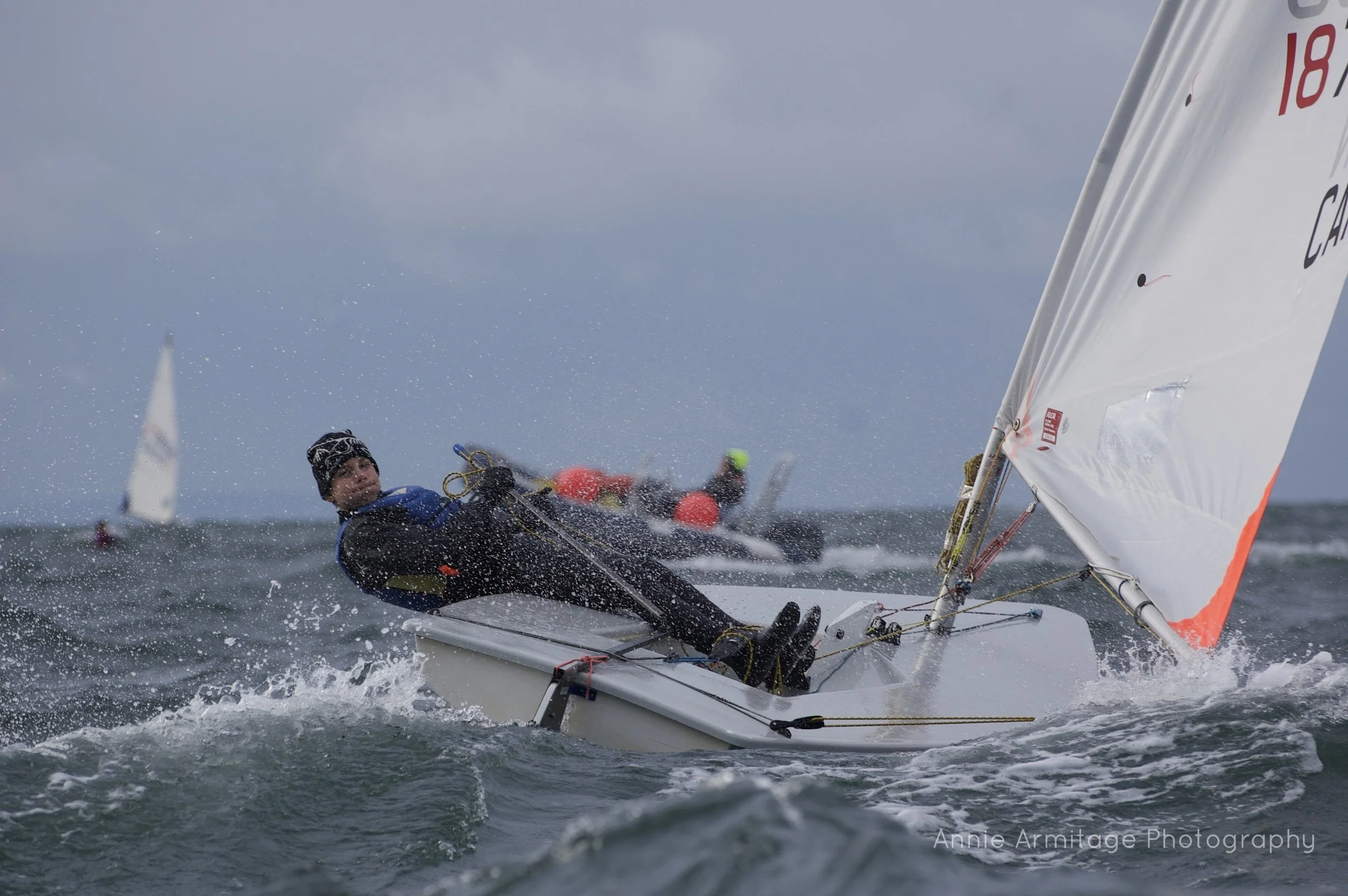 A person sailing a small sailboat in rough waters, wearing a black beanie and waterproof gear, with another sailor visible in the background. The sky is overcast.