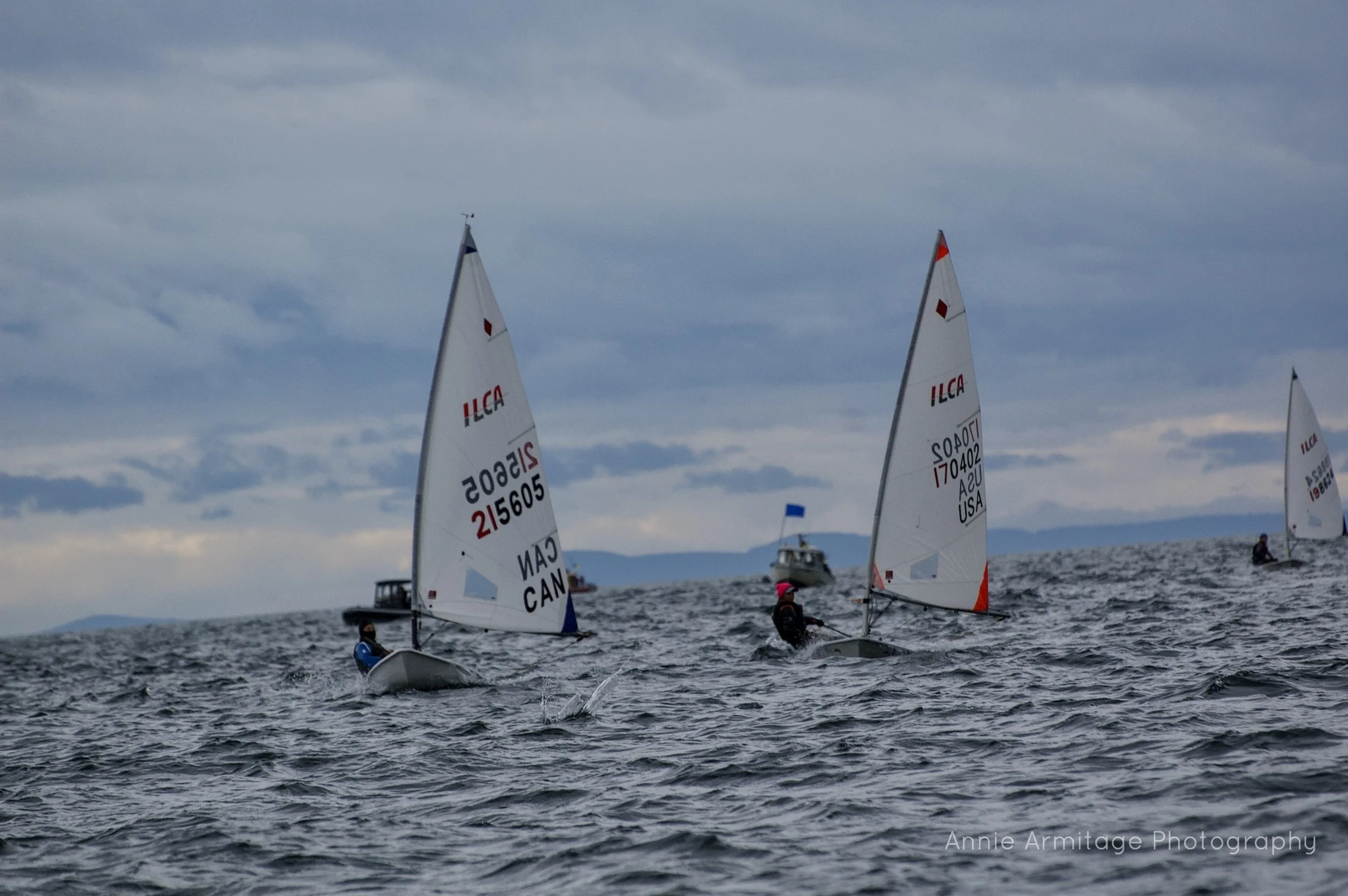 Several small sailboats with white sails racing on the water, under a cloudy sky, with a boat and distant land visible in the background.