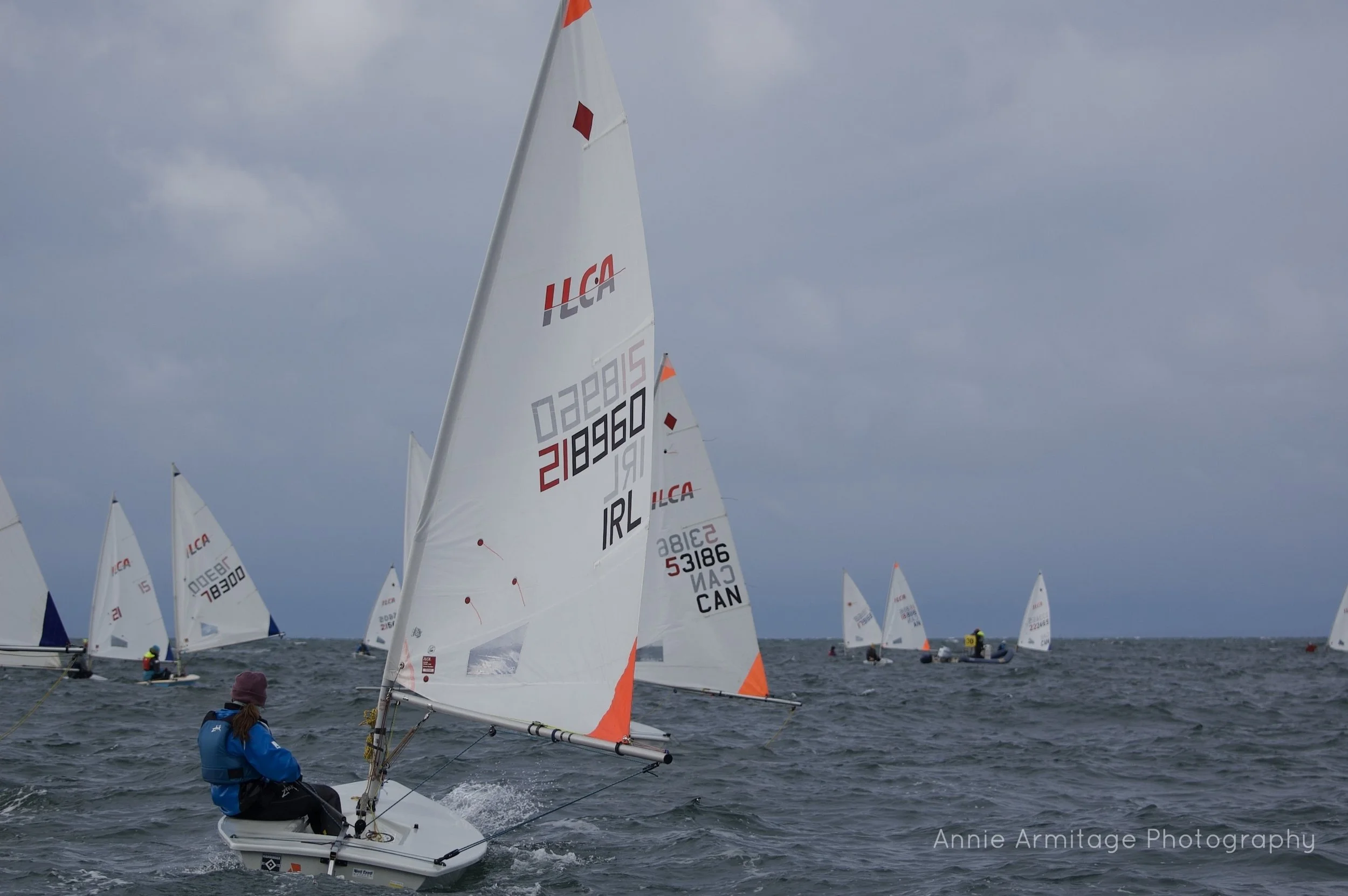 Multiple small sailboats racing on choppy water under cloudy sky, with a sailor in blue jacket and purple hat in the foreground, and the scene credited to Annie Armitage Photography.