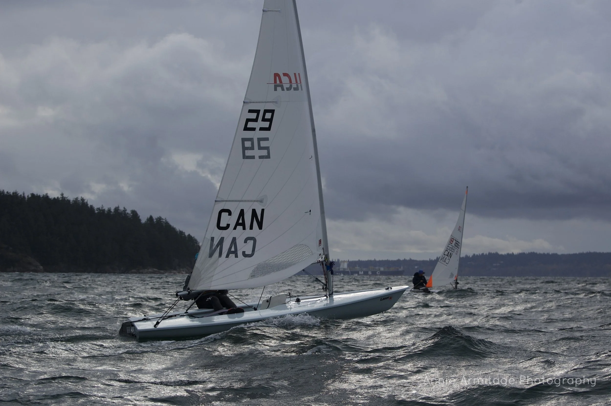 Two sailboats race on choppy water under a cloudy sky, with a distant shoreline and wooded hills in the background.