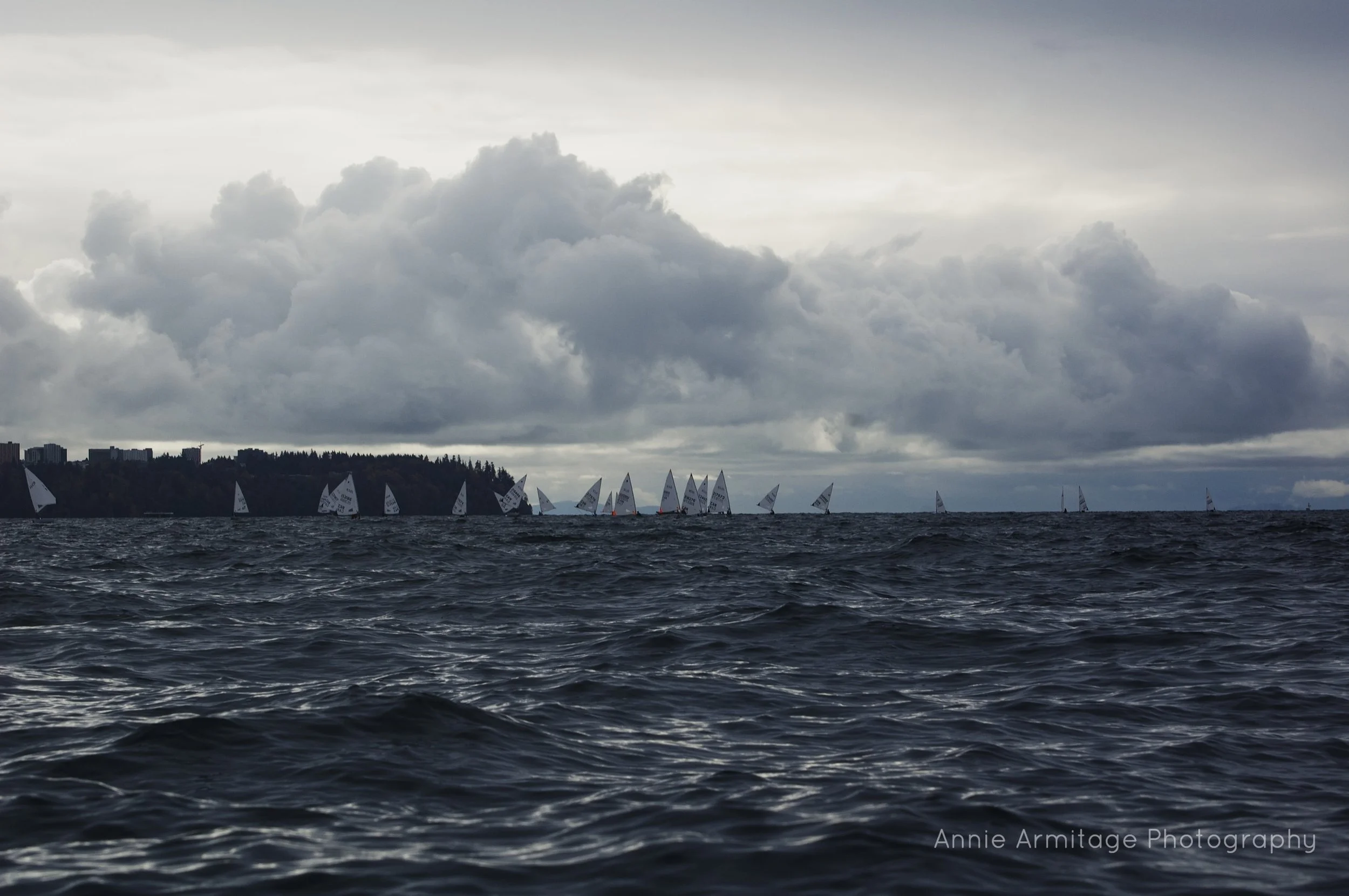 Overcast sky with dark clouds over a large body of water with several sailboats in the distance.