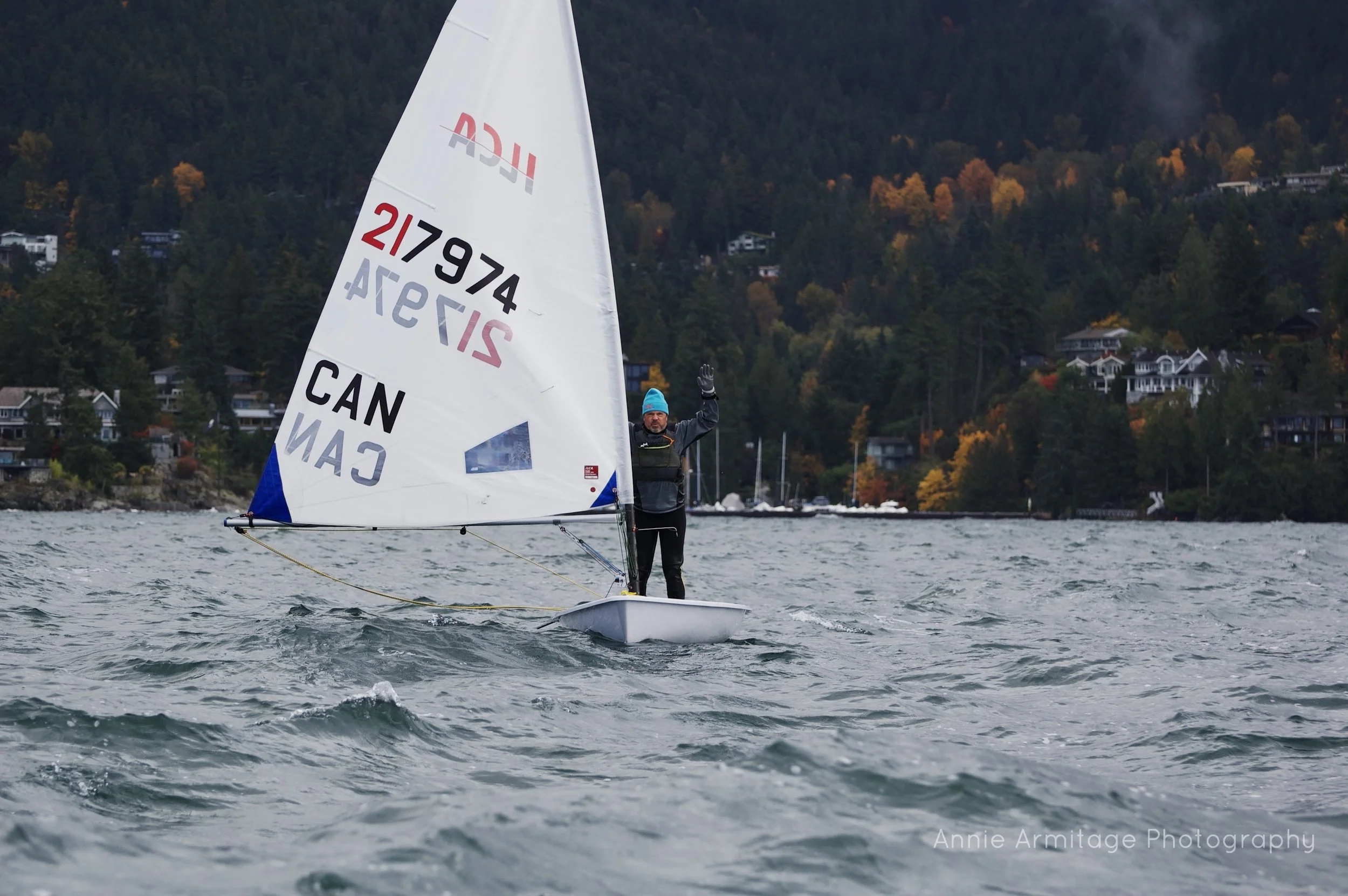 A person sailing a small boat on a body of water with a hilly area and houses in the background during overcast weather.