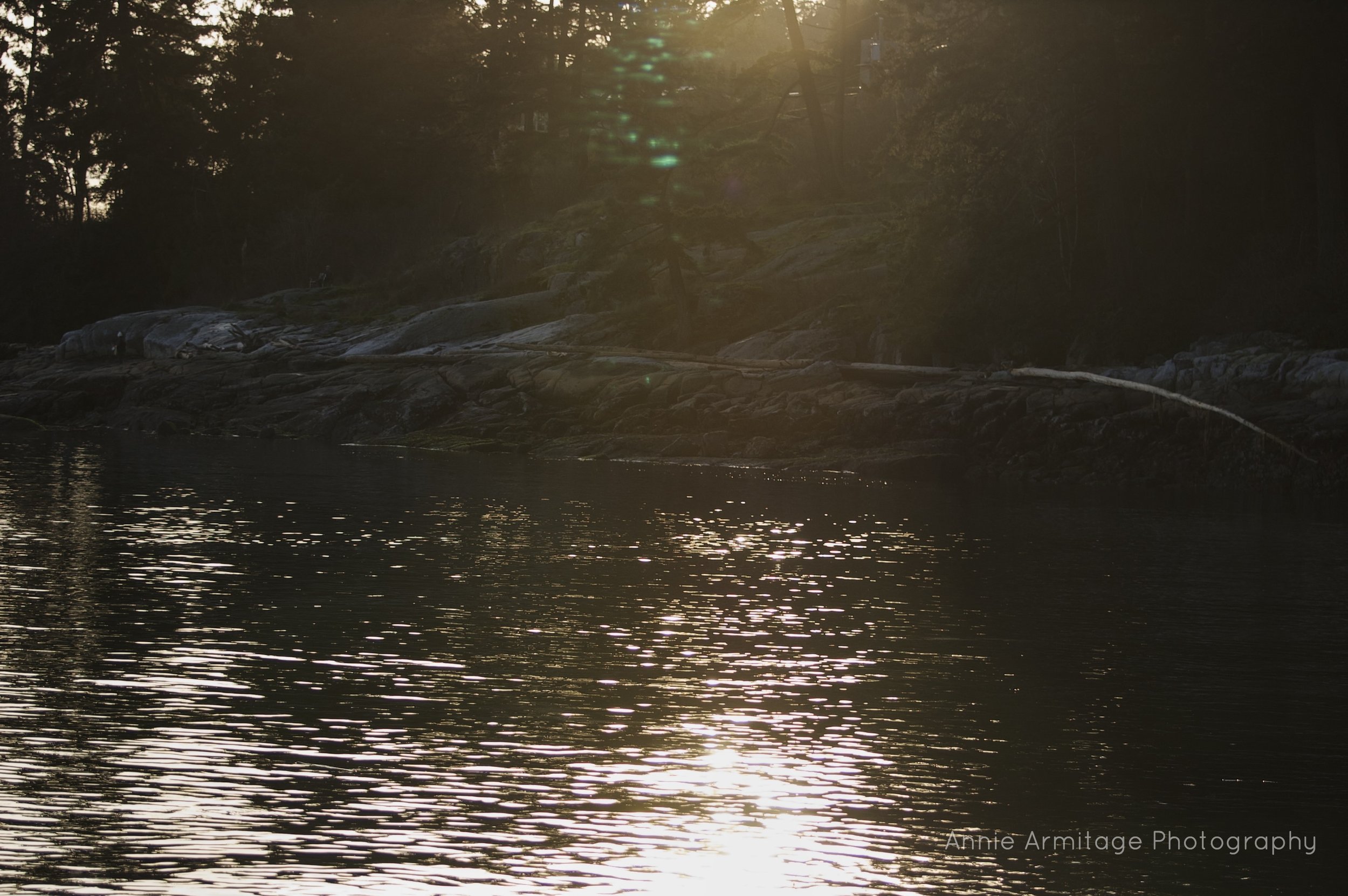 Sunset over a river with reflections, rocky shoreline, and trees in the background.