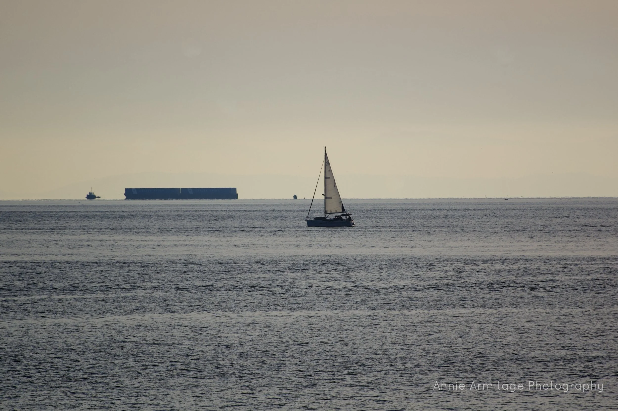 A sailboat on calm ocean waters with a cargo ship in the distance under a hazy sky.