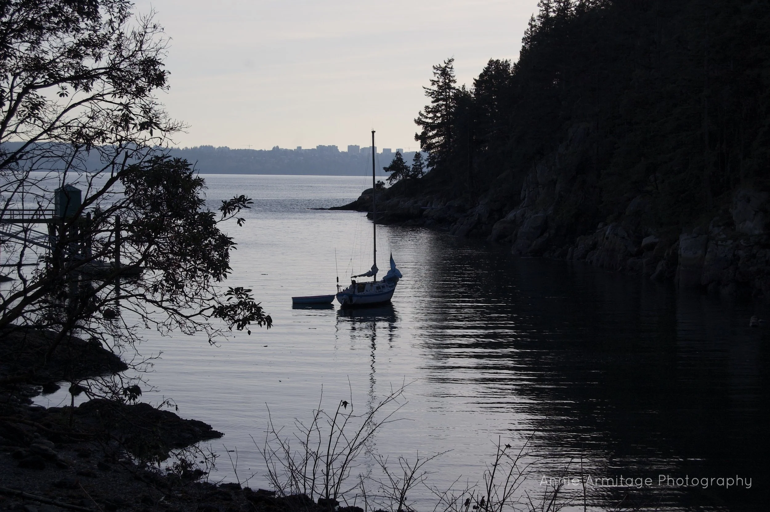A tranquil scene of a small sailboat and a rowboat anchored near a rocky shoreline with trees, overlooking a body of water and distant city skyline at sunset or dusk.