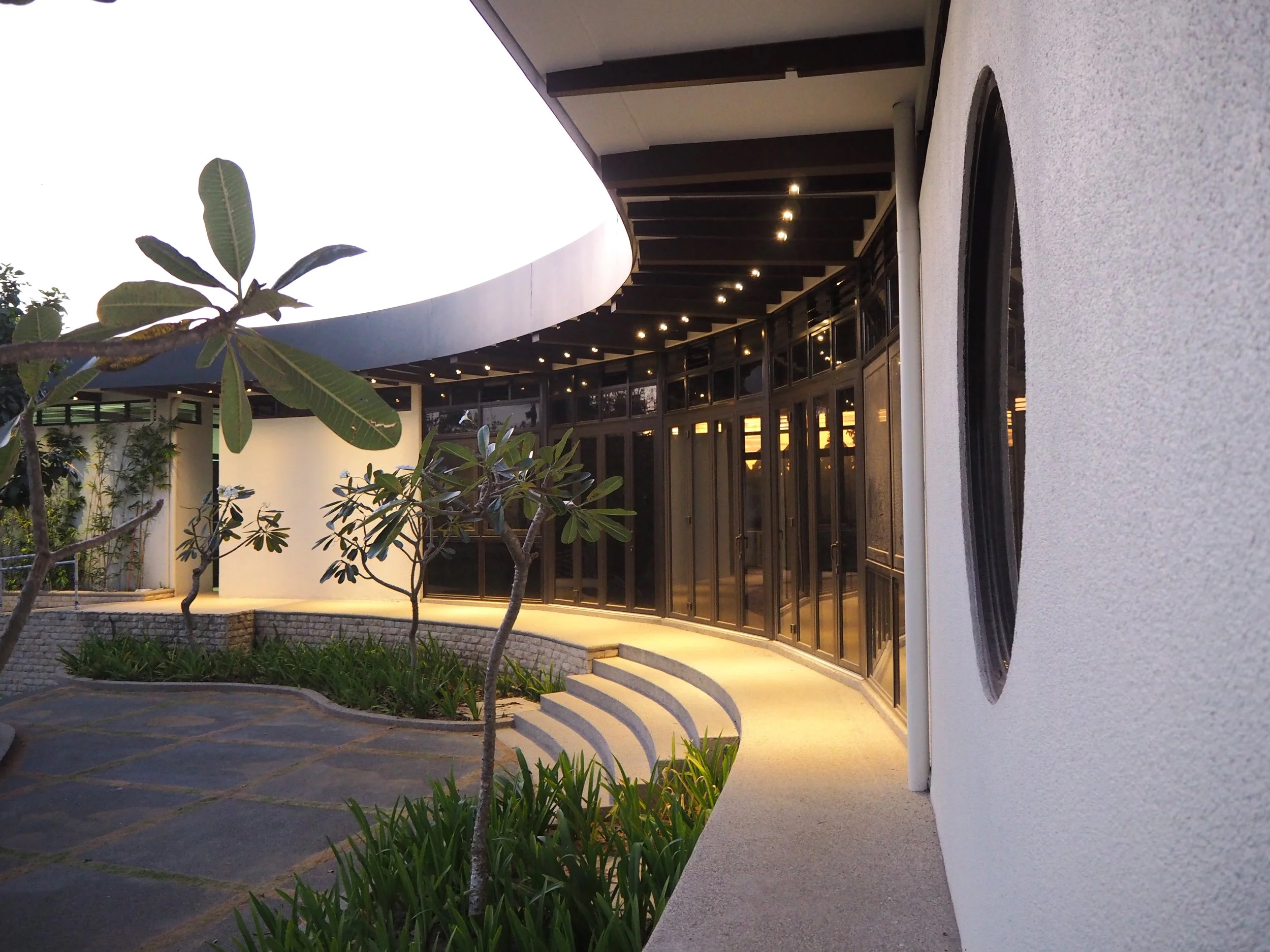 Exterior view of a modern building with curved design, glass windows, outdoor lighting, and landscaped garden with small trees and plants.