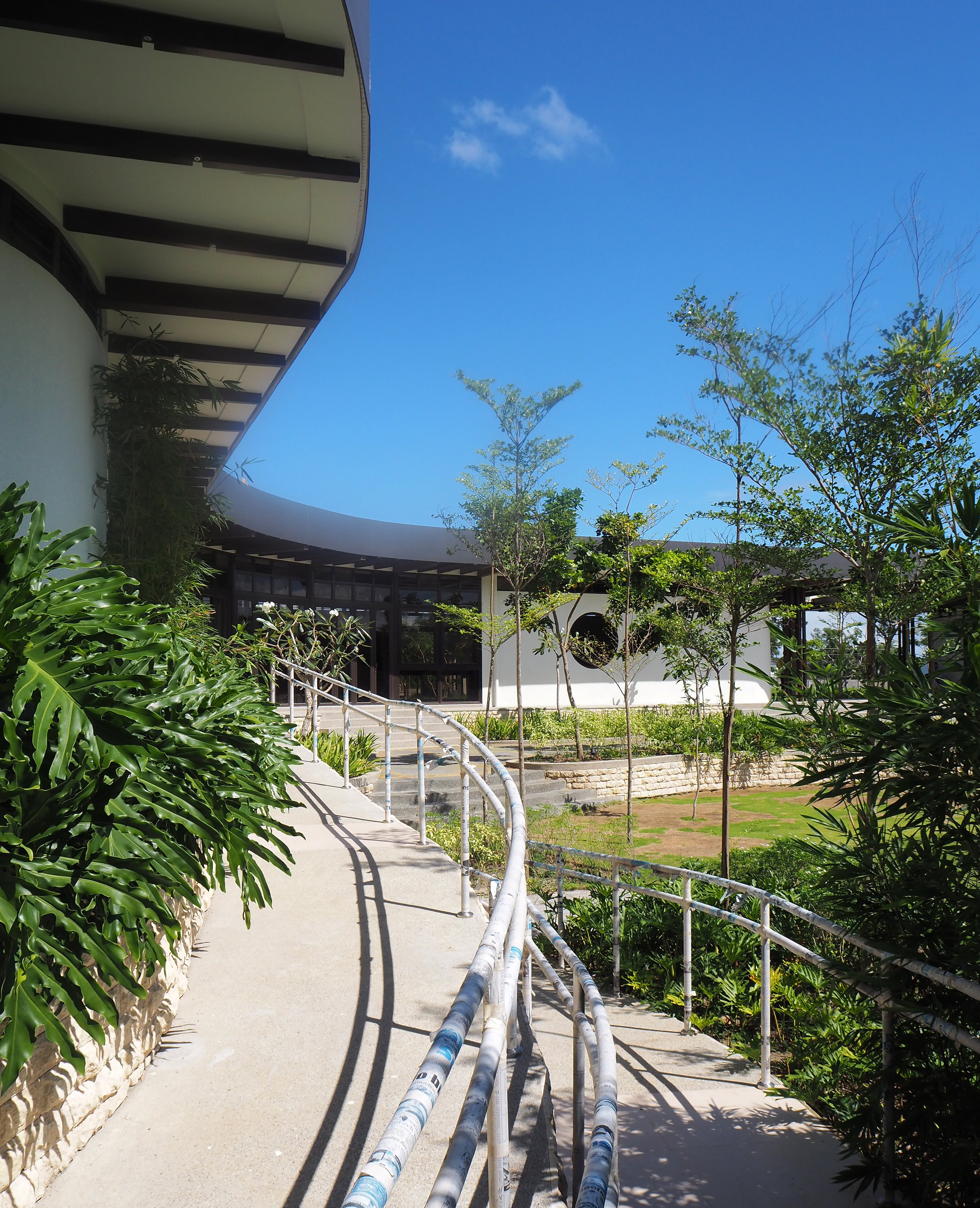 Curved walkway leading to a modern building with large windows and a white exterior, surrounded by trees and greenery under a clear blue sky.