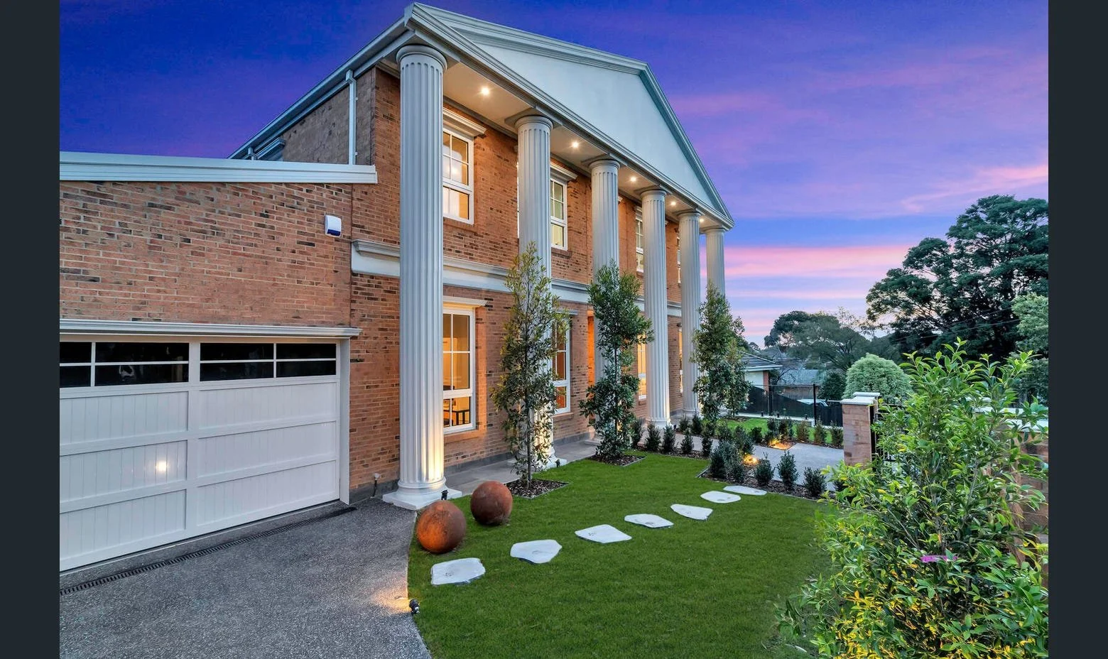 Exterior of a two-story brick house with large white pillars, illuminated windows, a white garage door, and a well-maintained lawn with stepping stones, small trees, and decorative spherical stones at dusk.