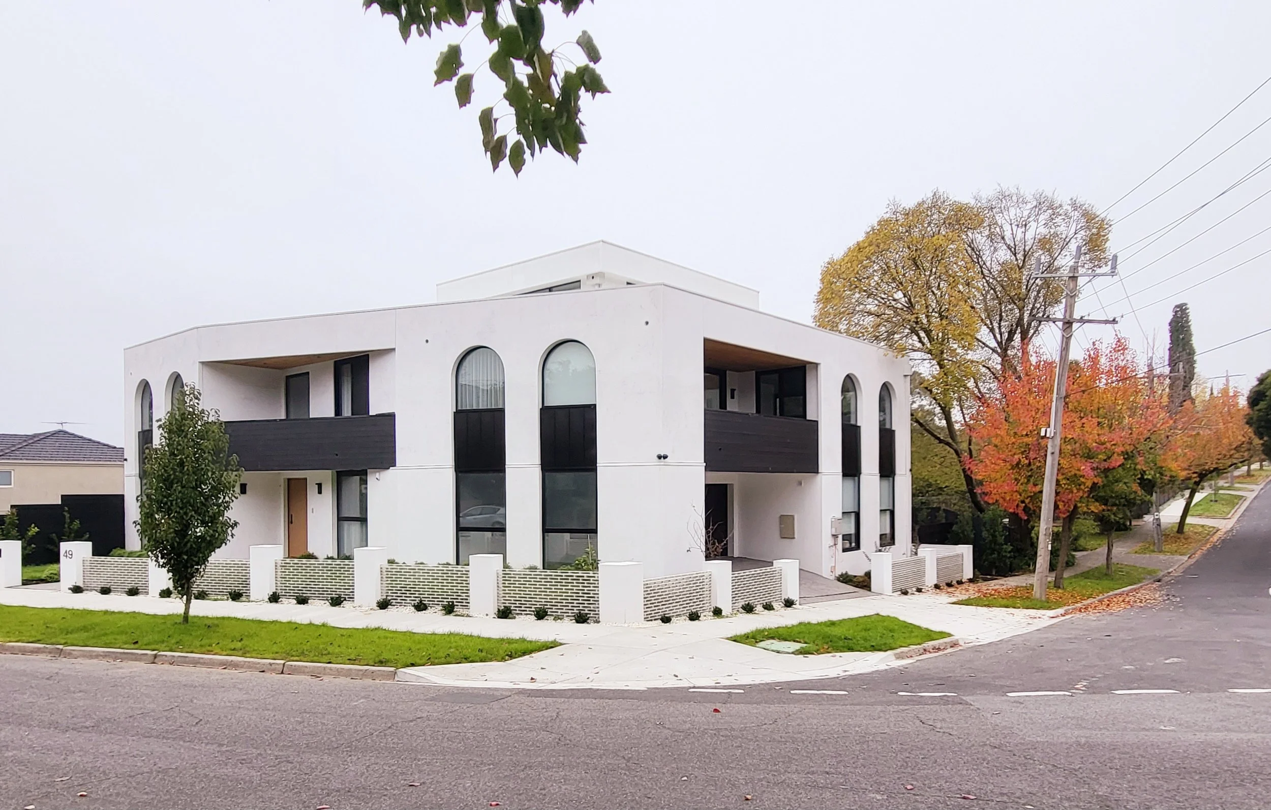 Modern white multi-story house with arched windows, black balconies, and a small front yard with a tree. The street has autumn trees with orange and yellow leaves, and a tilted utility pole on the sidewalk.