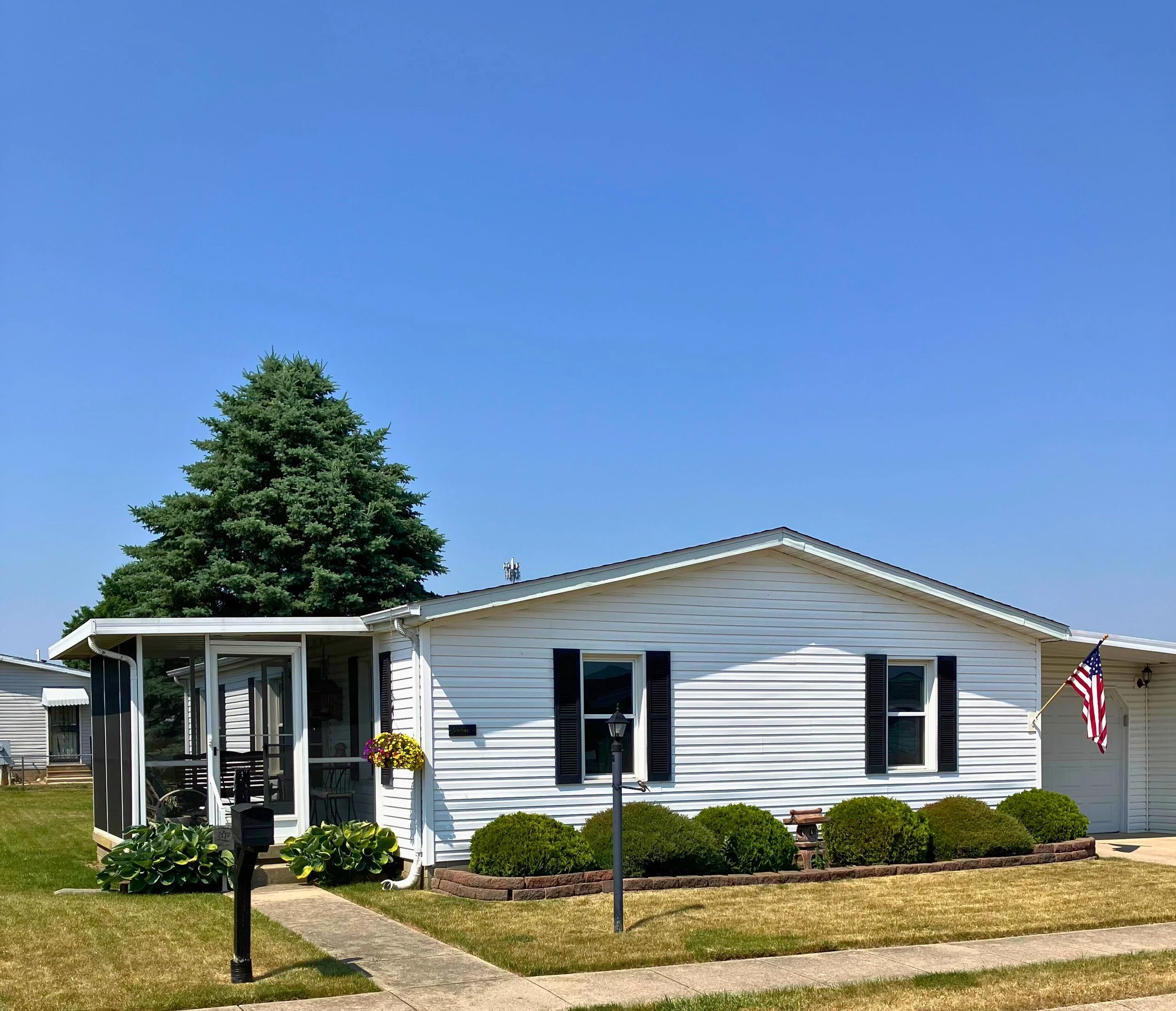 White house with black shutters, American flag, small front porch with hanging flower basket, next to large pine tree, set in a grassy lawn