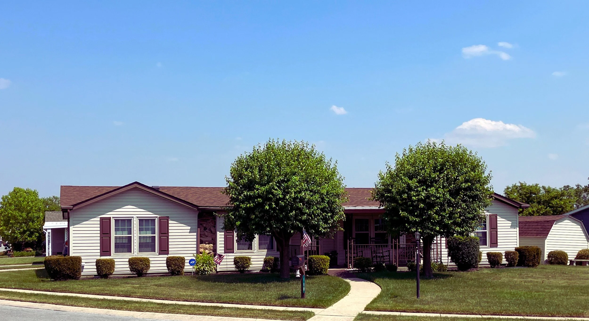 Single-story house with gray siding and purple shutters, two trees in front, manicured lawn, blue sky with few clouds.