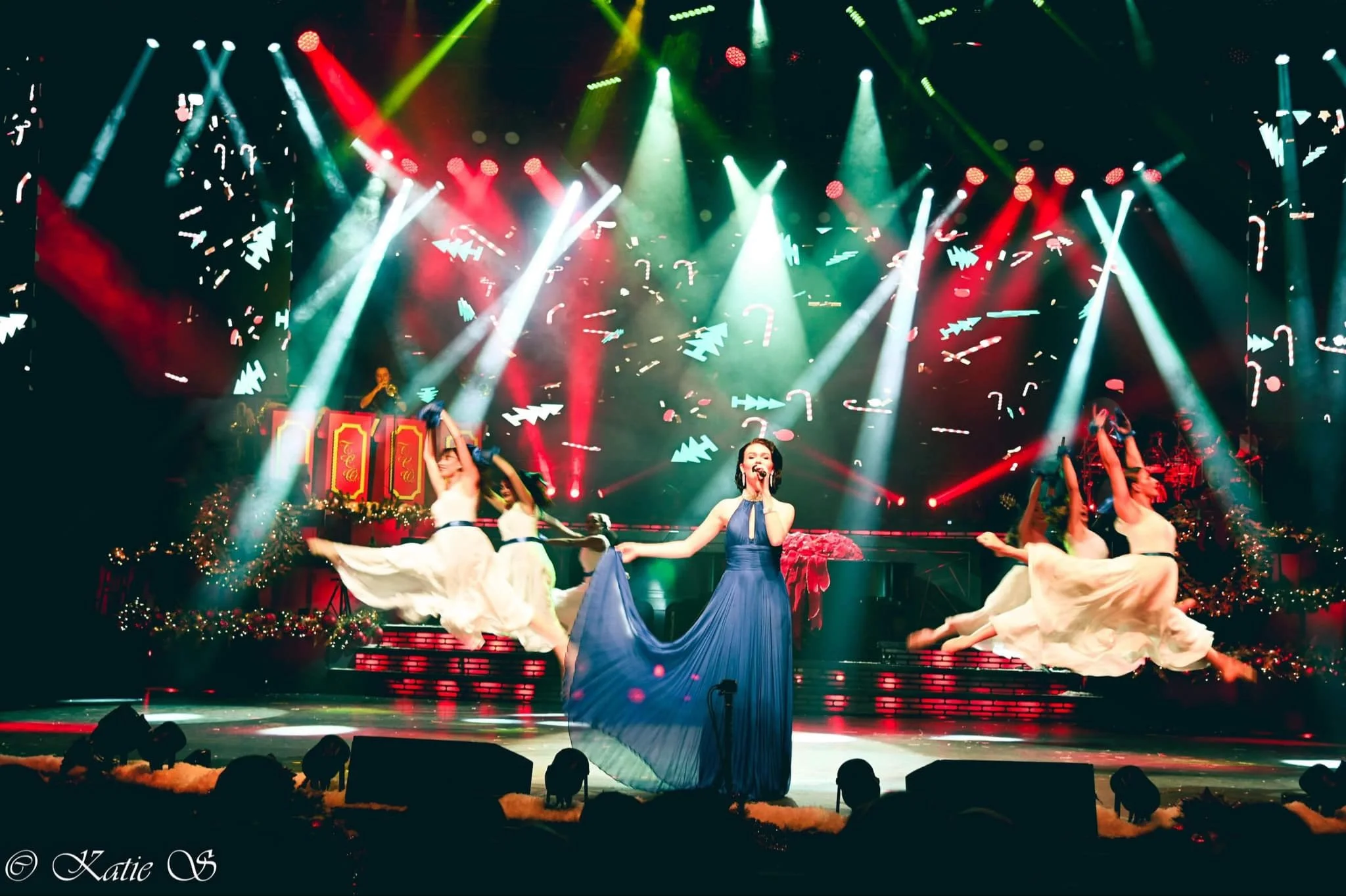 A Christmas-themed stage performance with a singer in a blue dress holding a microphone, surrounded by dancers in white dresses, with holiday decorations, Christmas trees, and colorful stage lights.
