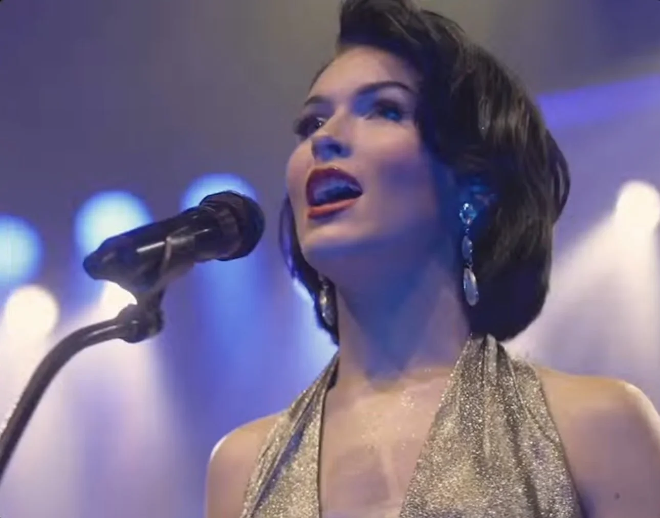 A woman singing into a microphone on stage, wearing a shimmering dress and large earrings with a blurred colorful background.