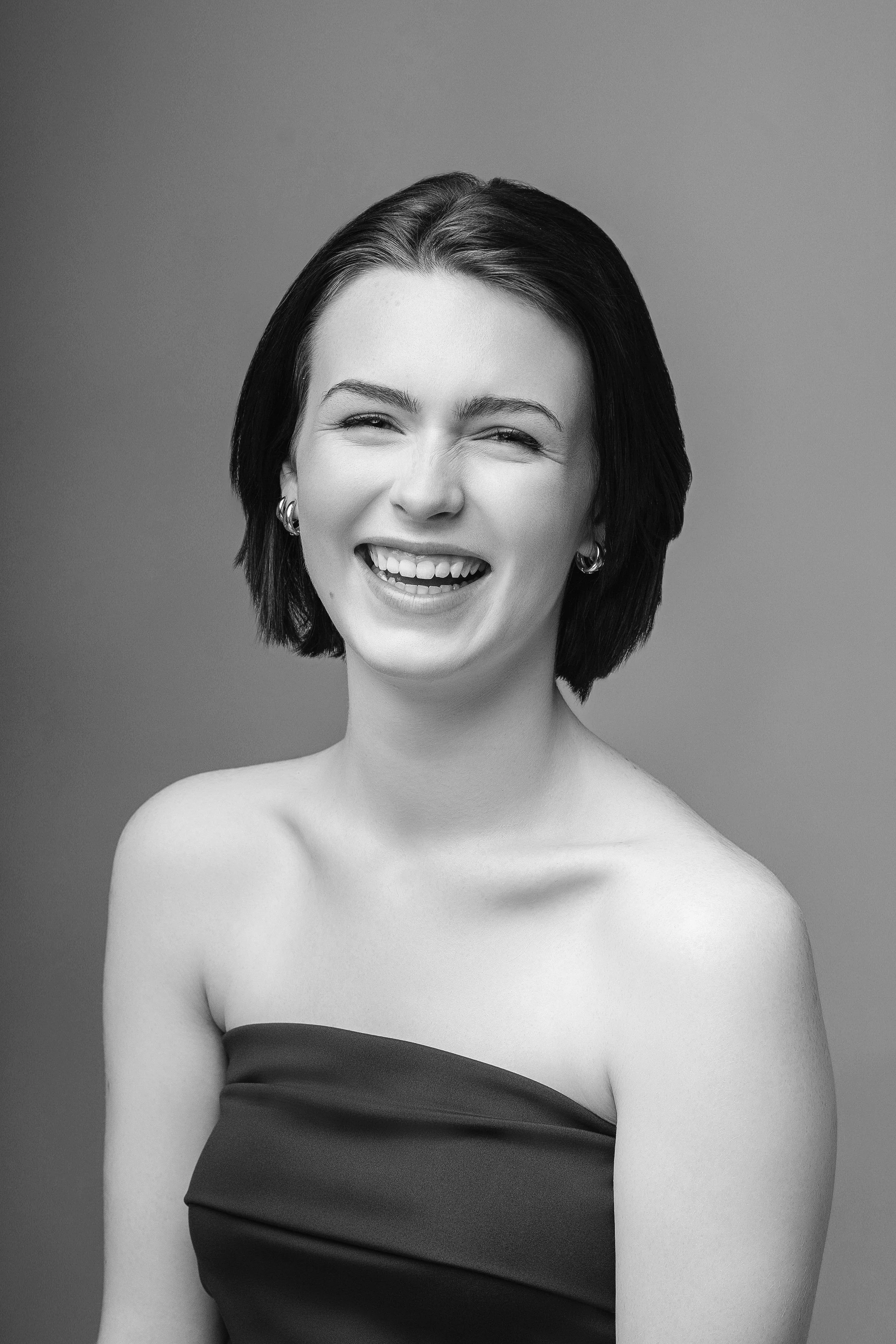 A black and white photo of a woman with short dark hair, smiling and winking, wearing a strapless top and hoop earrings.