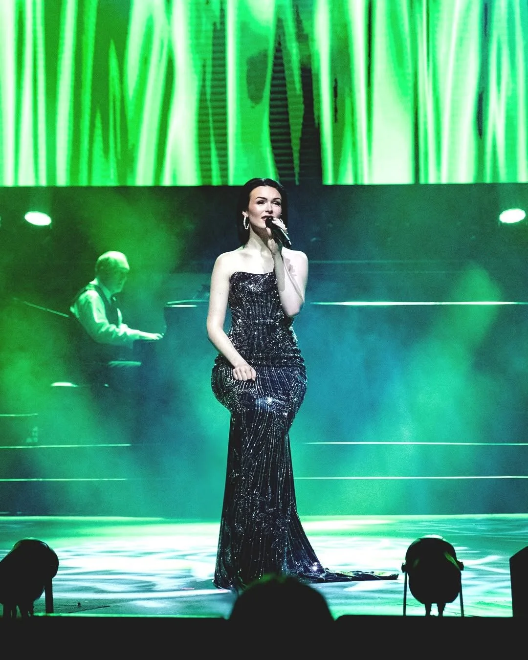 A singer in a black, glittery gown performing on stage with a pianist in the background, illuminated by green stage lights.