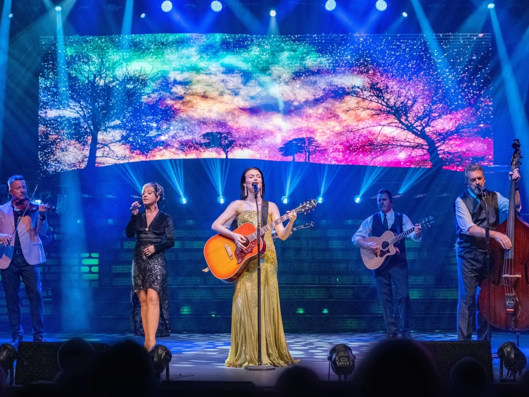 A musical band performing on stage with a colorful, starry backdrop and blue stage lighting.