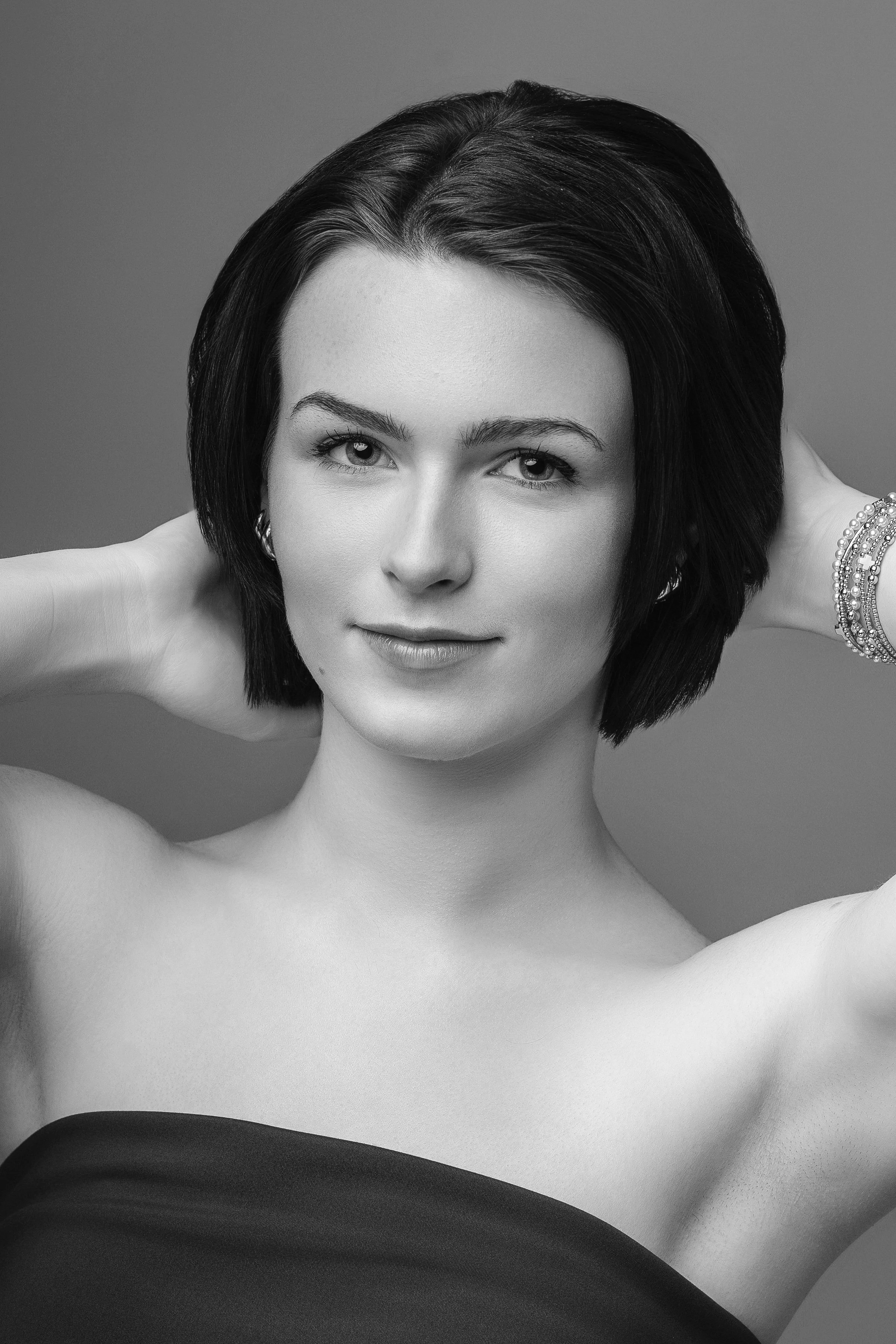 Black and white portrait of a woman with short dark hair, looking at the camera, smiling slightly, wearing a strapless top and jewelry, with her hands behind her head.