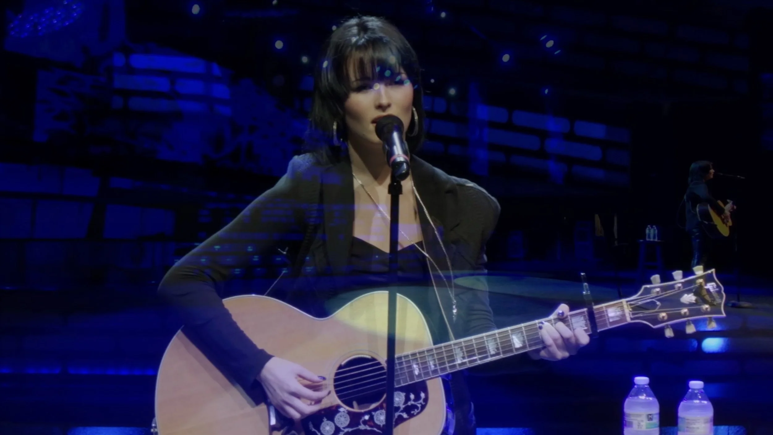 Woman with dark hair and wearing earrings playing an acoustic guitar and singing into a microphone on stage in a dimly lit venue.