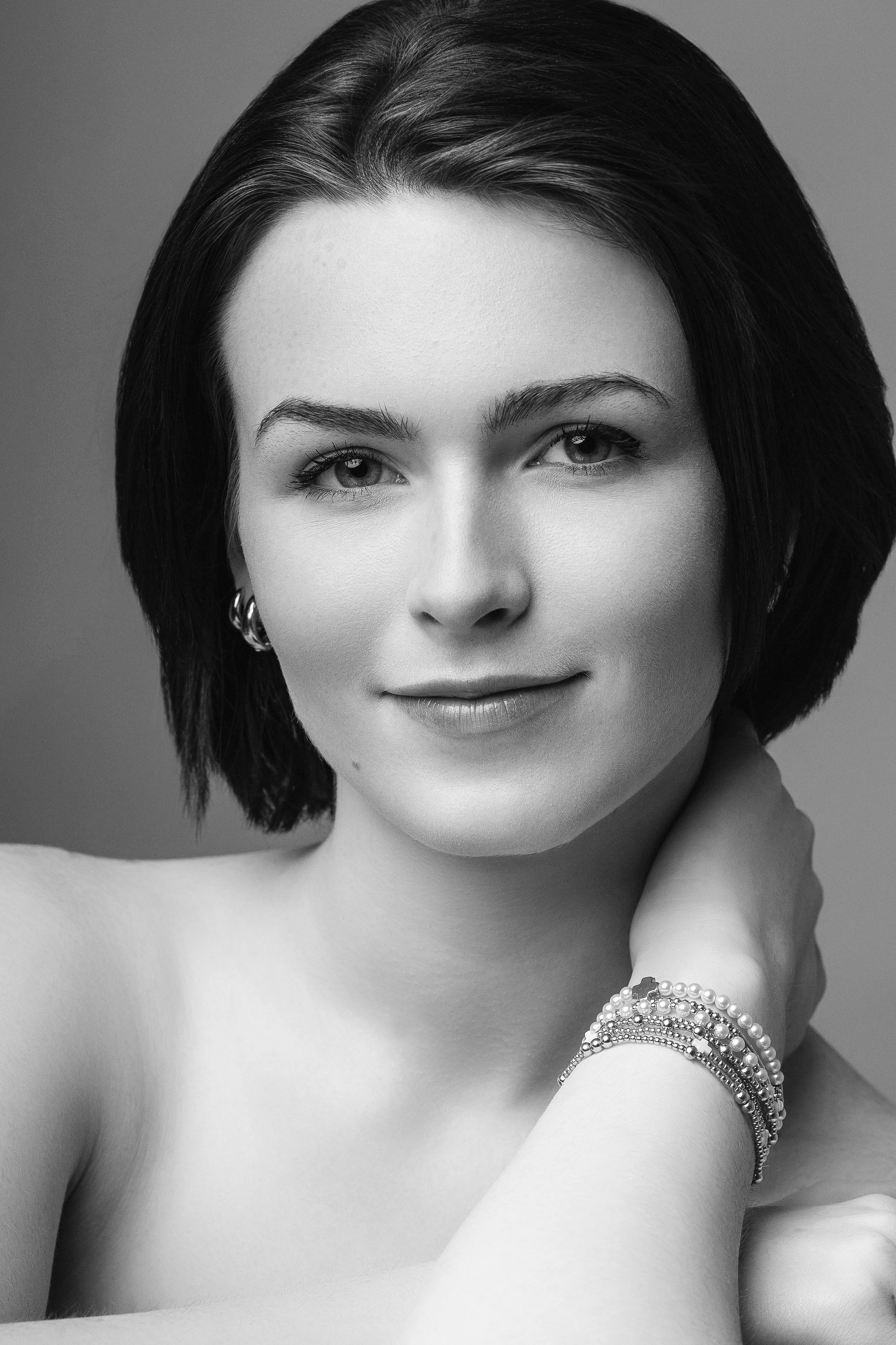 Close-up black-and-white portrait of a woman with dark hair, wearing pearl bracelets and earrings, smiling softly and looking at the camera, with one hand resting on her neck.