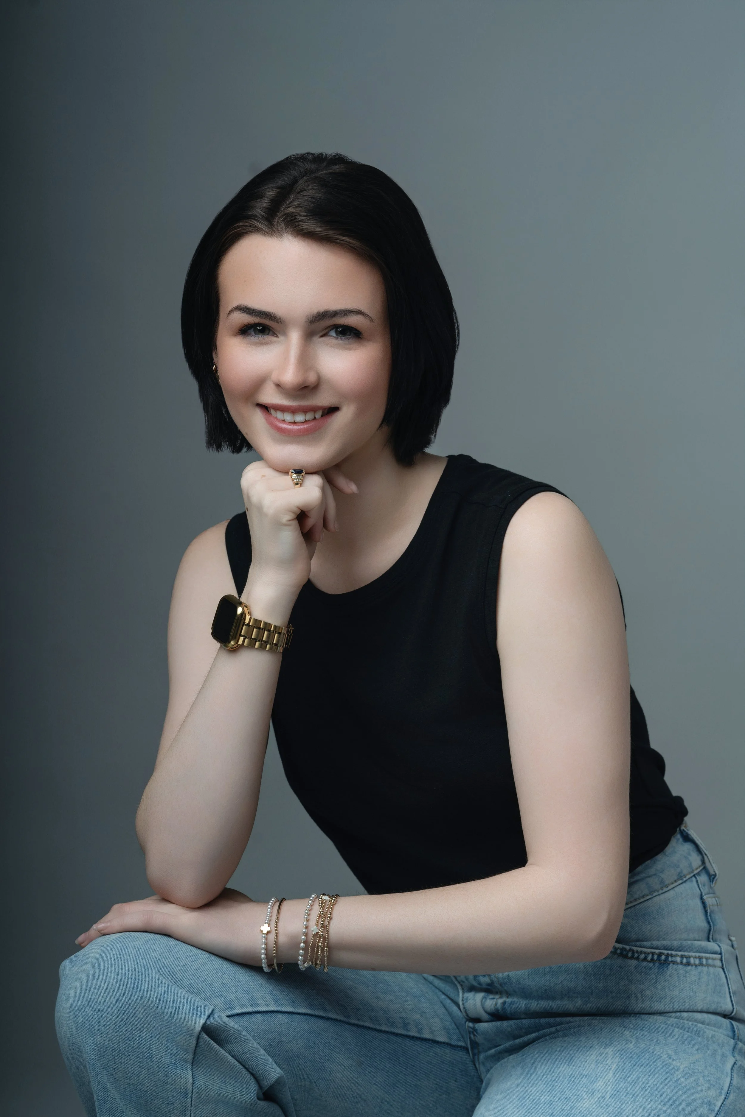 A young woman with short black hair, smiling, wearing a black sleeveless top, light blue jeans, a gold watch, and bracelets, sitting against a gray background.