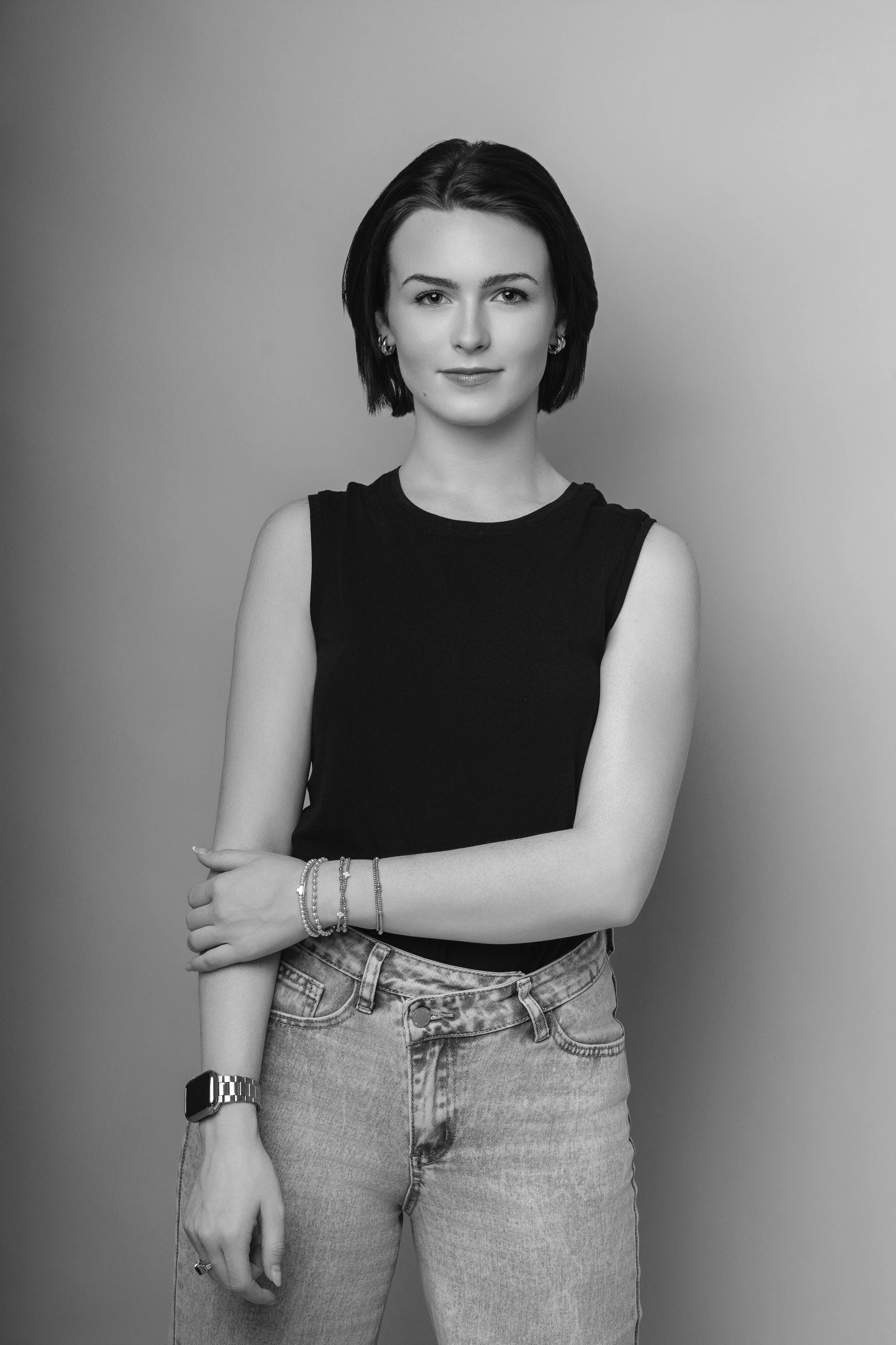Black and white photo of a young woman with short dark hair, wearing a sleeveless black top, light-colored jeans, and jewelry, standing against a plain wall.