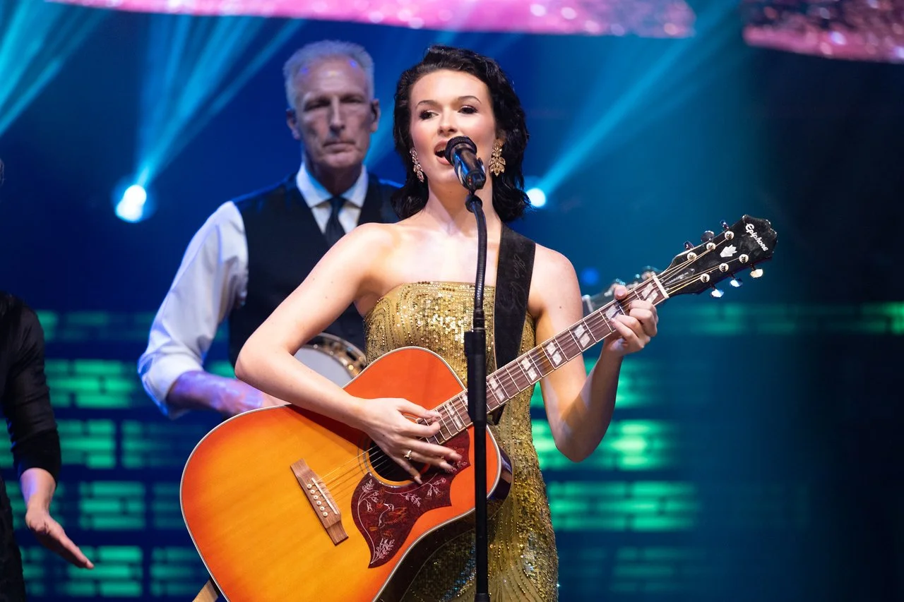 A woman with dark curly hair singing into a microphone while playing an orange acoustic guitar. She wears a gold dress and gold earrings. A man with gray hair, dressed in a black vest and white shirt, stands behind her, with colorful stage lights in 