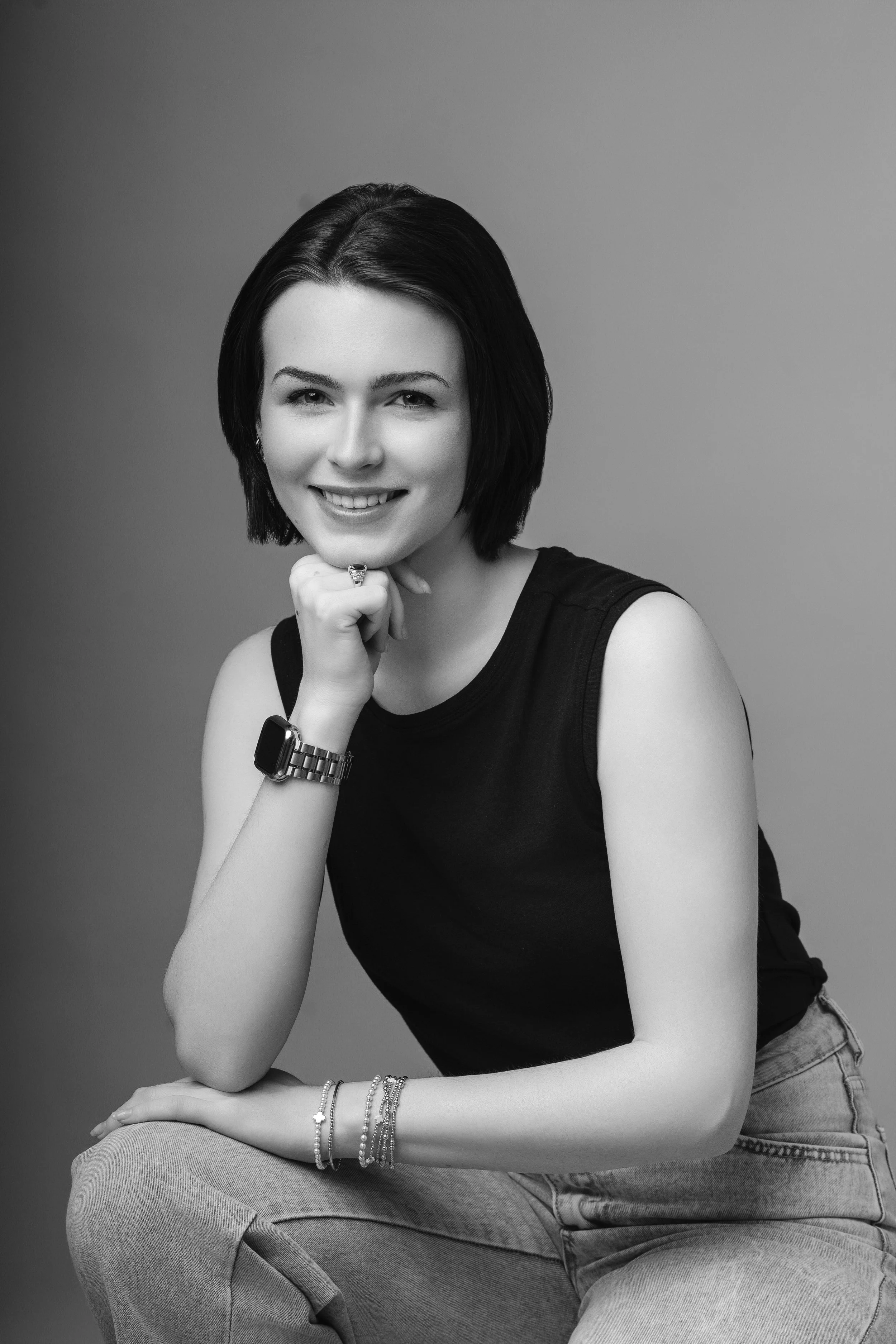 Black and white portrait of a young woman with short dark hair, wearing a sleeveless top, jeans, and jewelry, smiling with her chin resting on her hand.