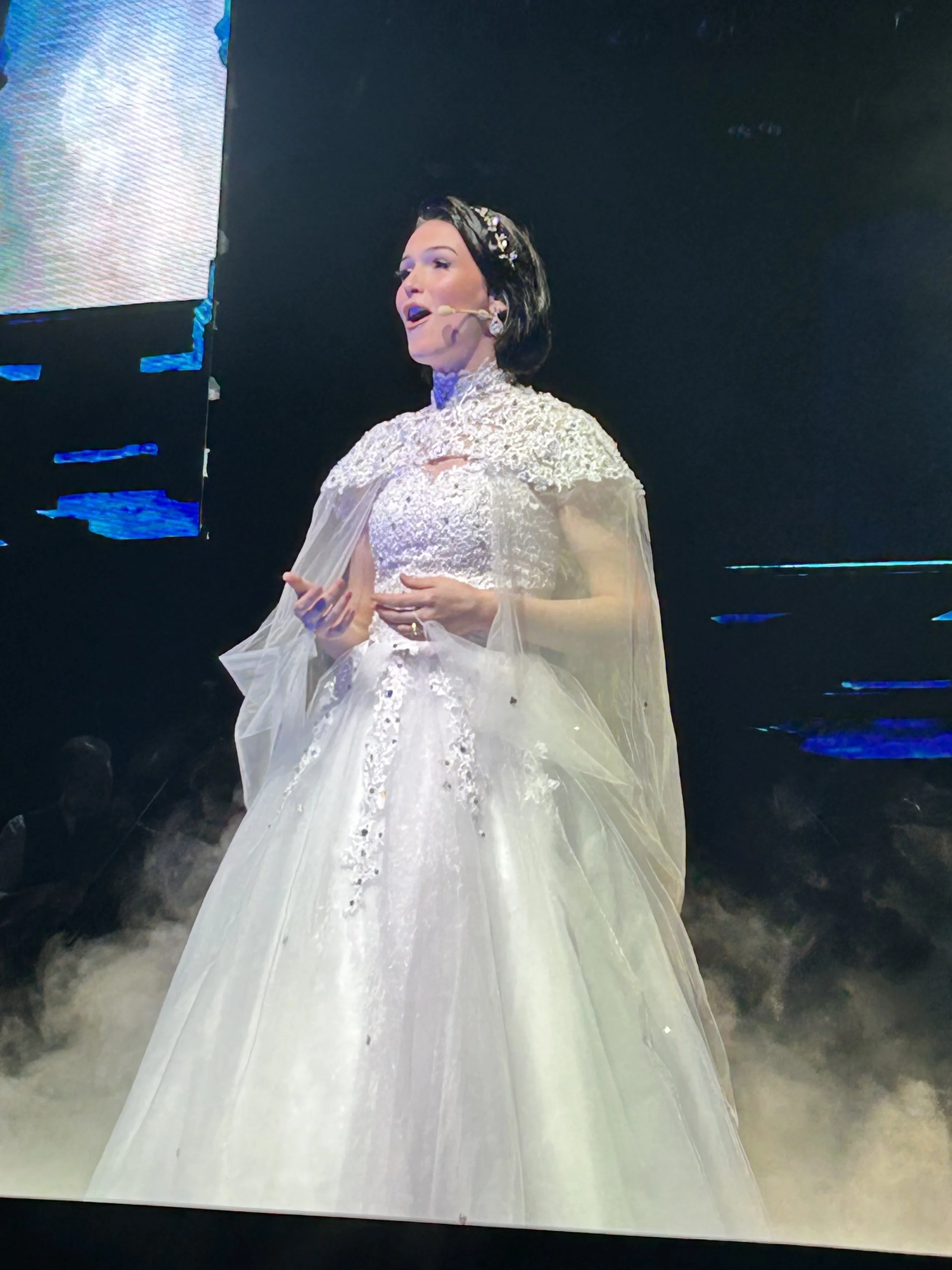 A woman dressed in a wedding gown and cape, speaking on stage with a headset microphone, dark background, and blue lighting effects.