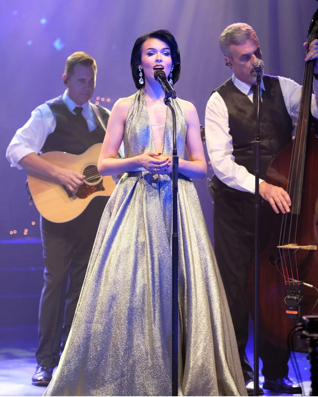A woman in a shiny gold gown singing into a microphone on stage, accompanied by two men playing guitar and double bass in the background.