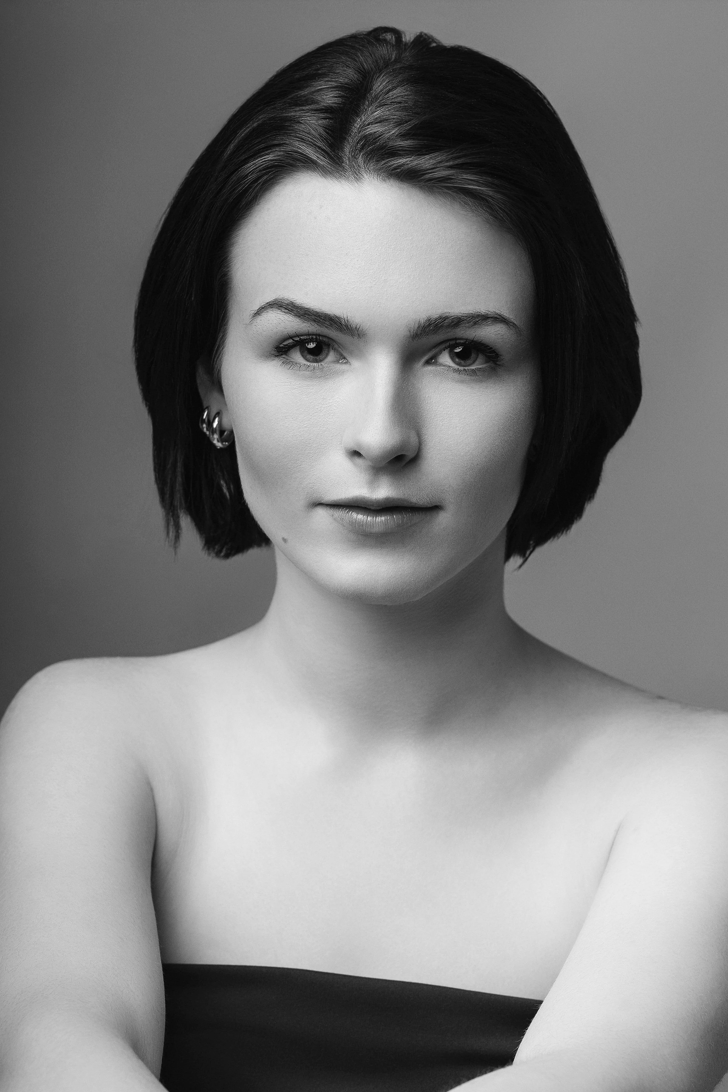 A black and white portrait of a young woman with short, dark hair, wearing earrings, looking directly at the camera, with a neutral expression.