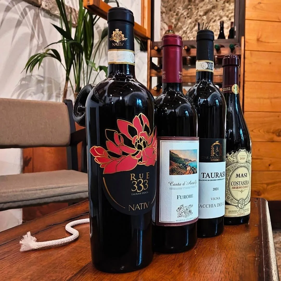 Quattro bottiglie di vino sul tavolo in una stanza con pareti in legno e decorazioni in piante in background.