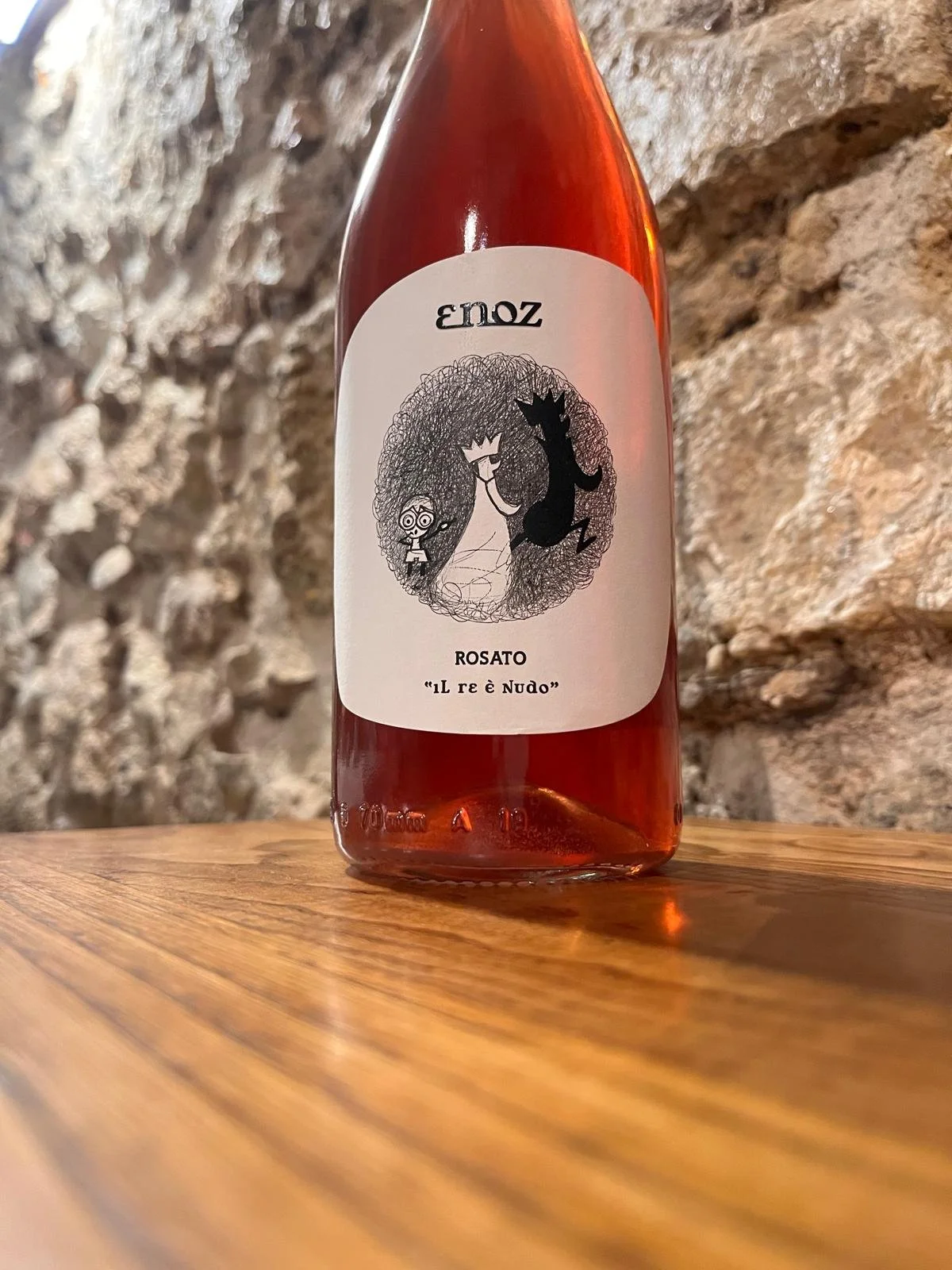 Bottiglia di vino rosato con etichetta disegnata, posizionata su un tavolo di legno con sfondo di muro di pietra.