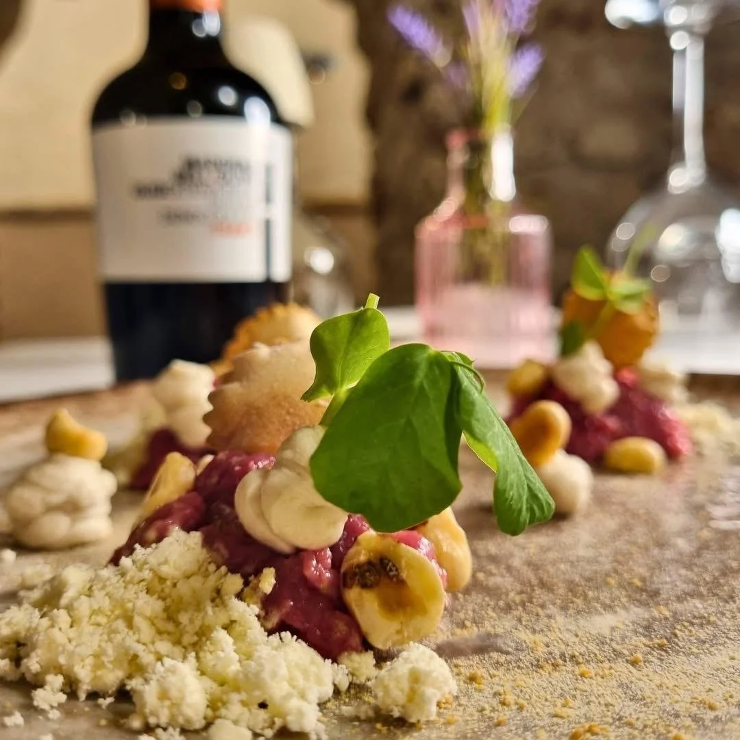 Distribuzione di un piatto di pasta con crema colorata, noci, e decorato con una foglia verde, su un piano di legno, con bottiglia di vino e vaso con fiori sullo sfondo.