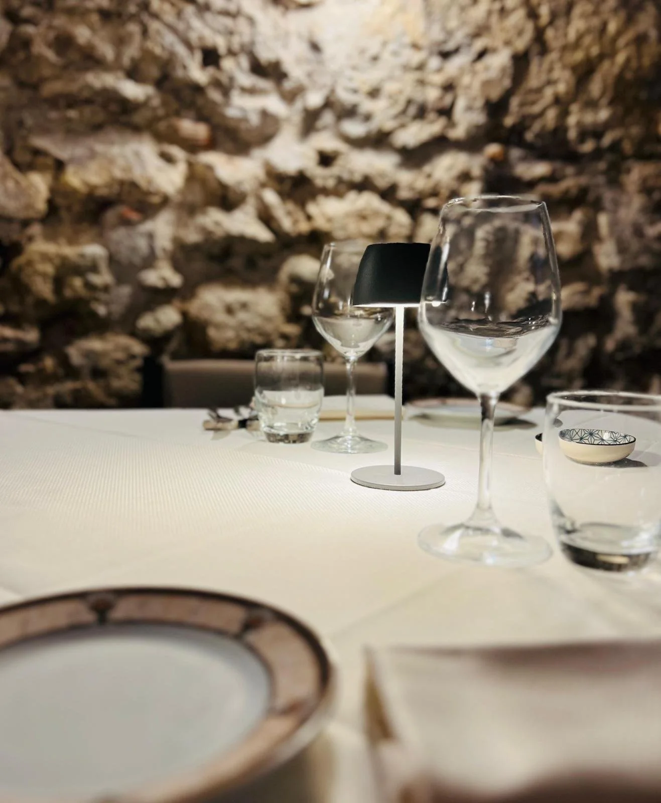 Tavola apparecchiata con bicchieri, piatti e un lampadario al centro, con uno sfondo di parete in pietra rustica.