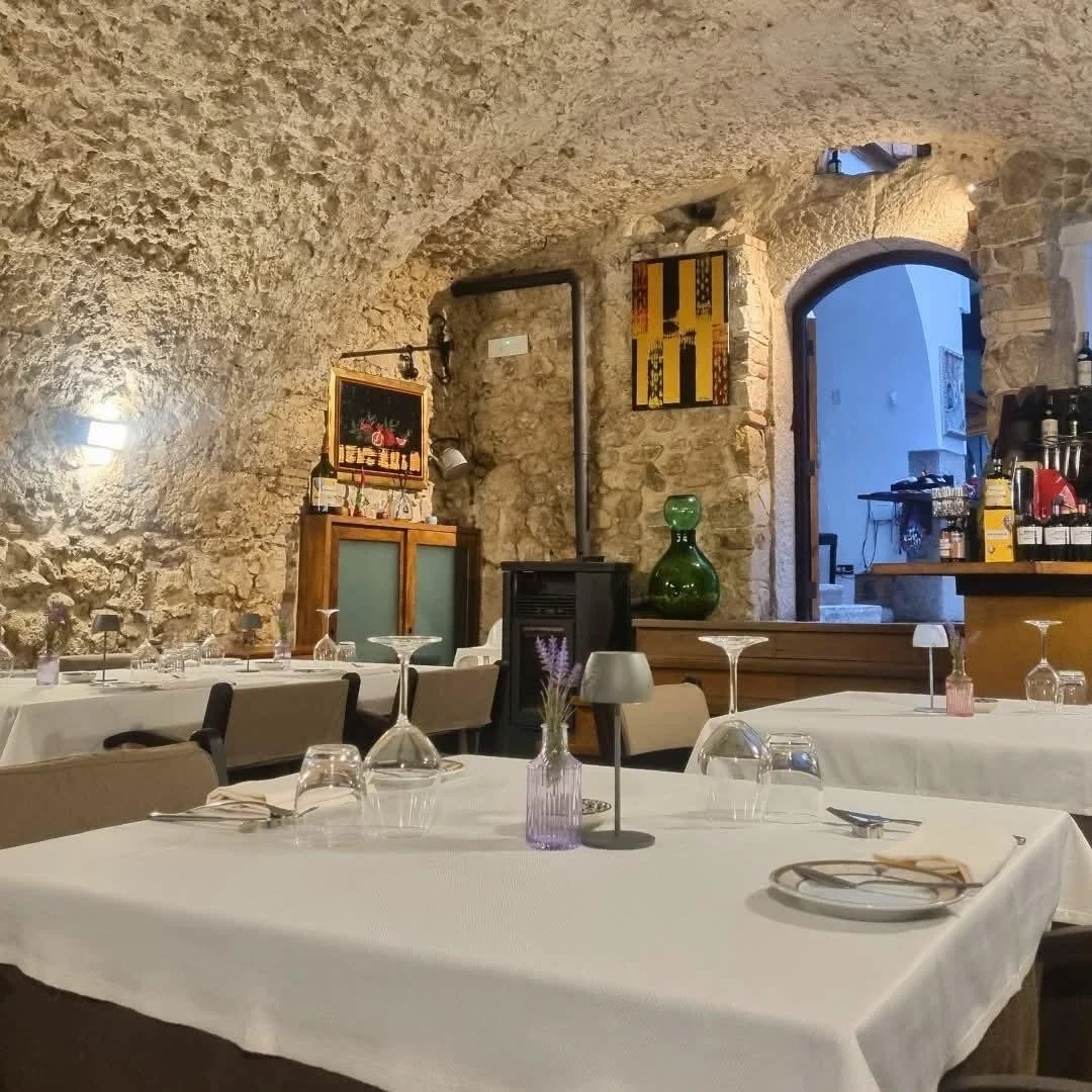 Interno di un ristorante con pareti di pietra, tavoli con tovaglie bianche, bicchieri capovolti e decorazioni floreali, illuminazione calda e un arco in pietra che conduce a una zona con pareti in pietra e decorazioni.