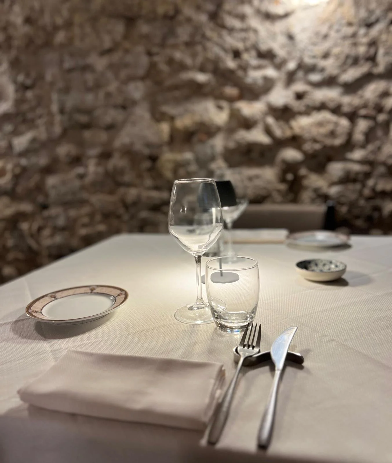 Tavola apparecchiata con piatti, bicchieri, posate, e tovaglioli, con uno sfondo di muro di pietra in un ristorante elegante.