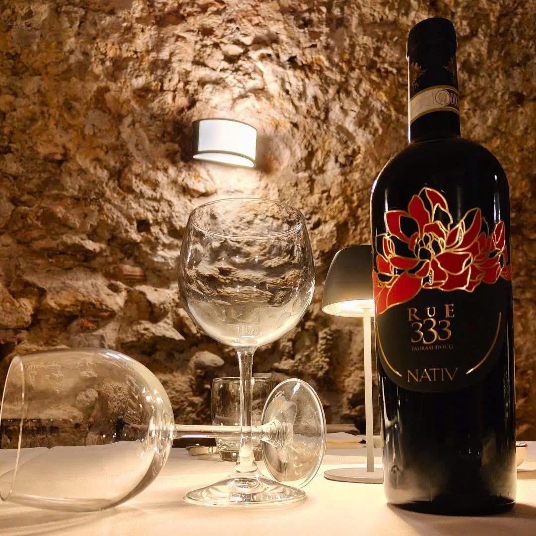 Una bottiglia di vino rosso con etichetta nera e rossa, due bicchieri da vino, uno rovesciato e uno in piedi, e una lampada vicino a una parete di pietra, in un ambiente interno.