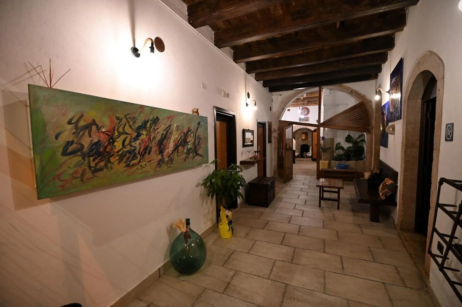 Interno di un hotel rustico con pareti bianche, soffitto in legno e arredi in stile tradizionale. Decorazioni artistiche alle pareti, piante e mobili in legno lungo il corridoio.