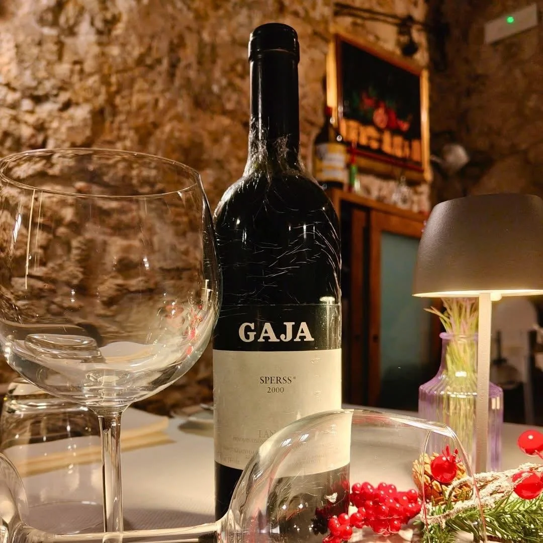 Bottiglia di vino Gaja con due bicchieri di vino vuoti, decorazioni di bacche rosse e un vaso con un fiore, ambiente accogliente con pareti di pietra e un quadro sullo sfondo.