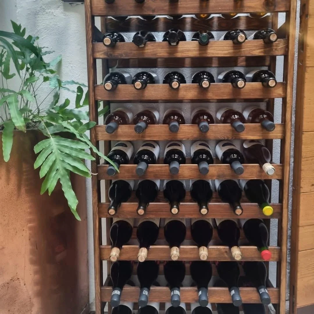 Scaffale in legno con bottiglie di vino posizionate orizzontalmente, alcune con etichette visibili, su un lato una pianta verde