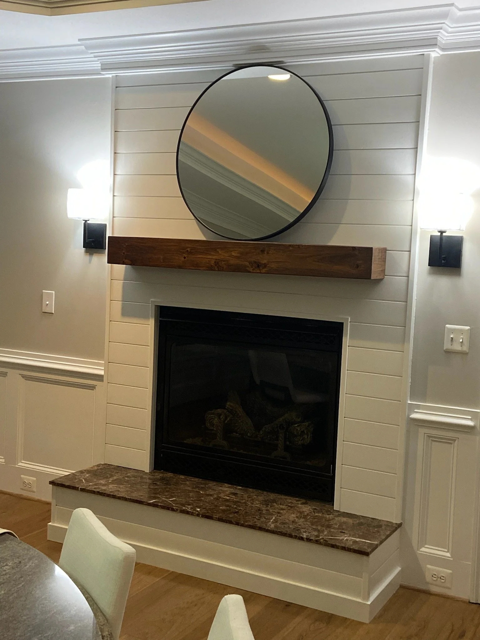 Living room fireplace with a black frame, a marble hearth, a wooden mantel, an oval mirror above the mantel, and white paneled walls with wall sconces.